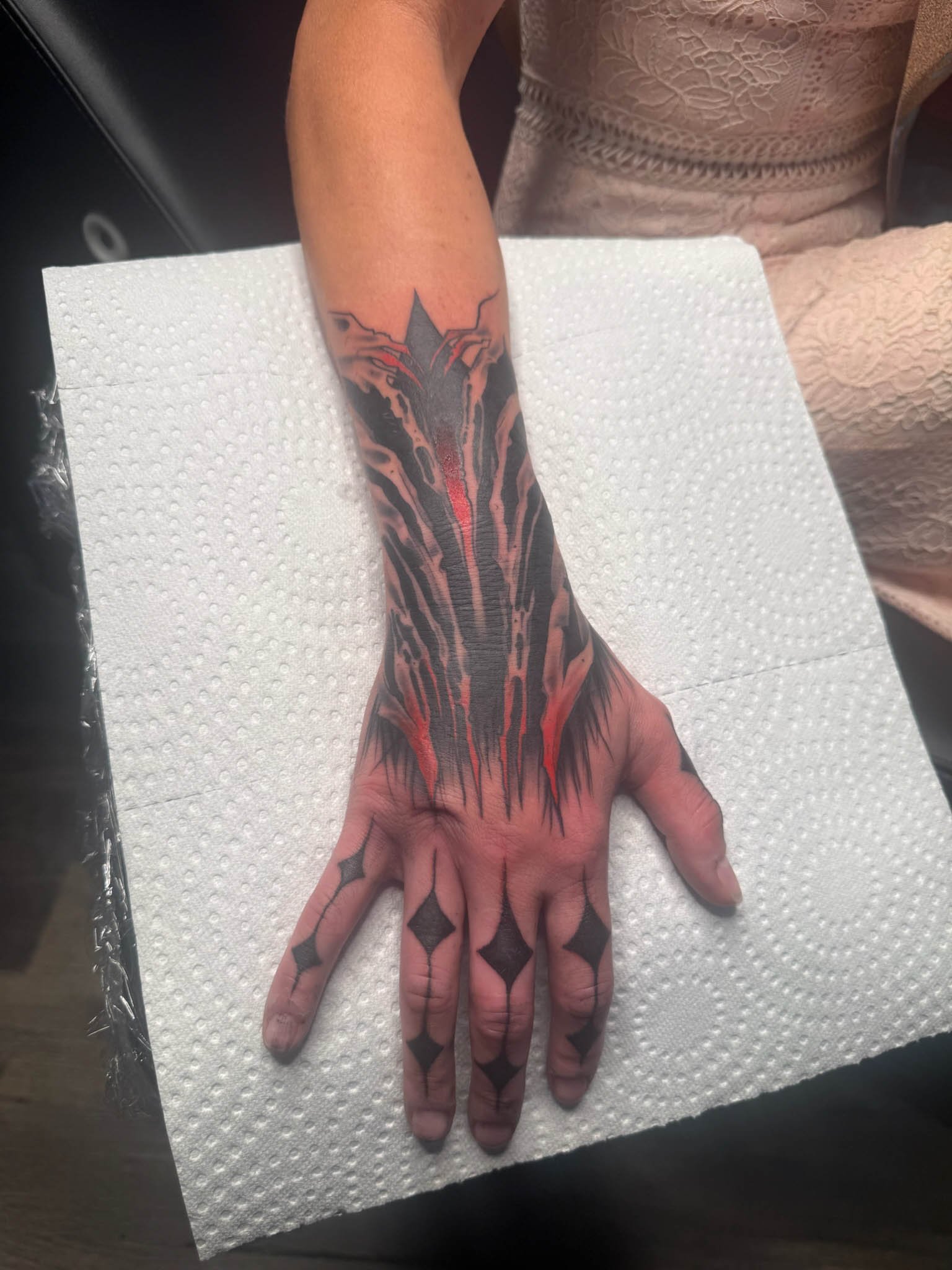 Tattoo of a black and red dragon on a person's forearm and hand, with black diamond shapes on fingers, on a white paper towel.