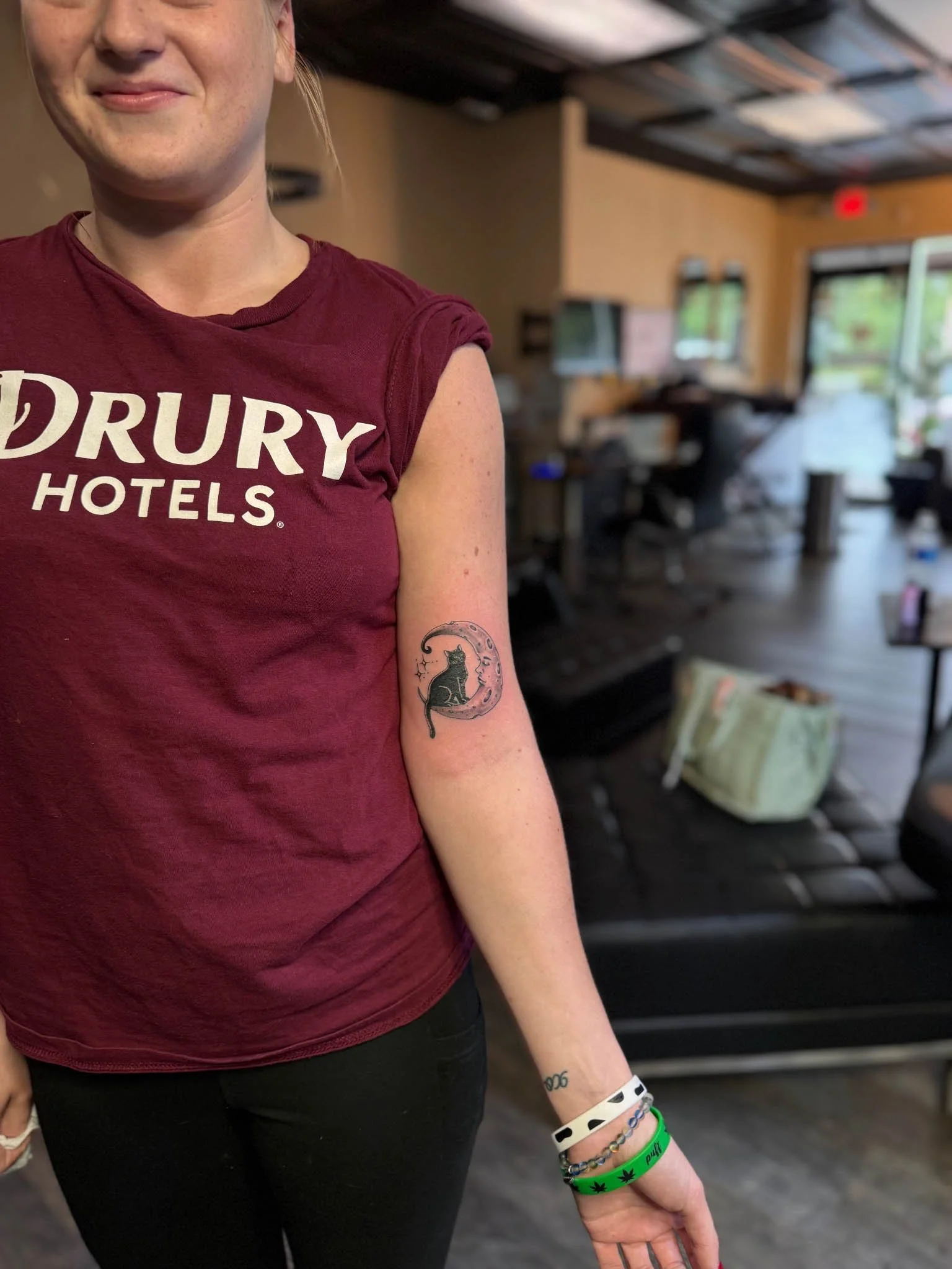 Alt Text: Small minimalist tattoo of a black cat silhouette sitting inside a decorative crescent moon with small star accents, located on the back of a person's upper arm