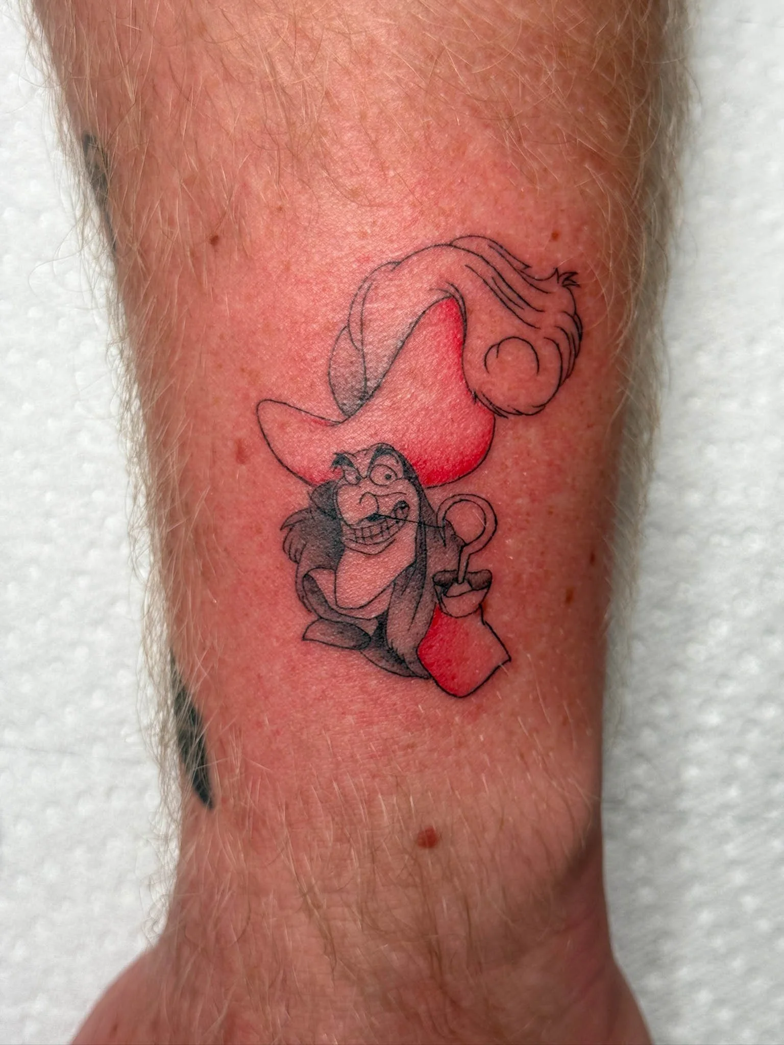 Tattoo of a cartoon witch with a big hat, squatting and holding a small bag, outlined in black with red shading.