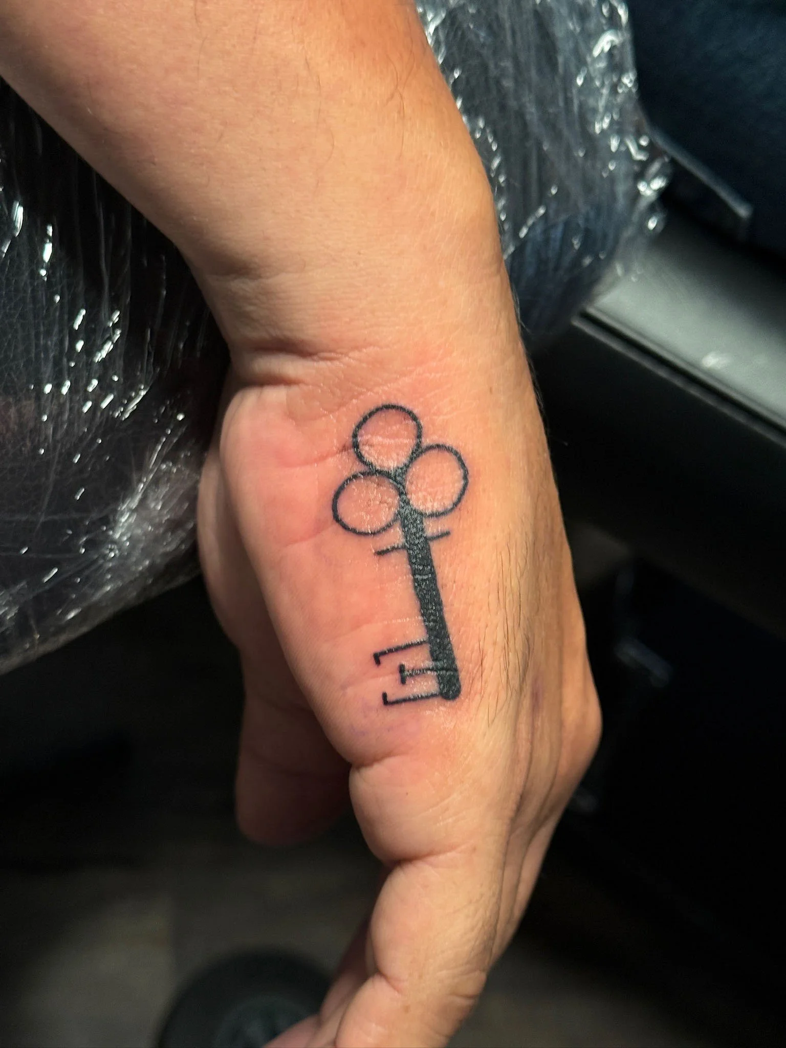 Tattoo of a simple black key with three circles on top, on the person's finger.