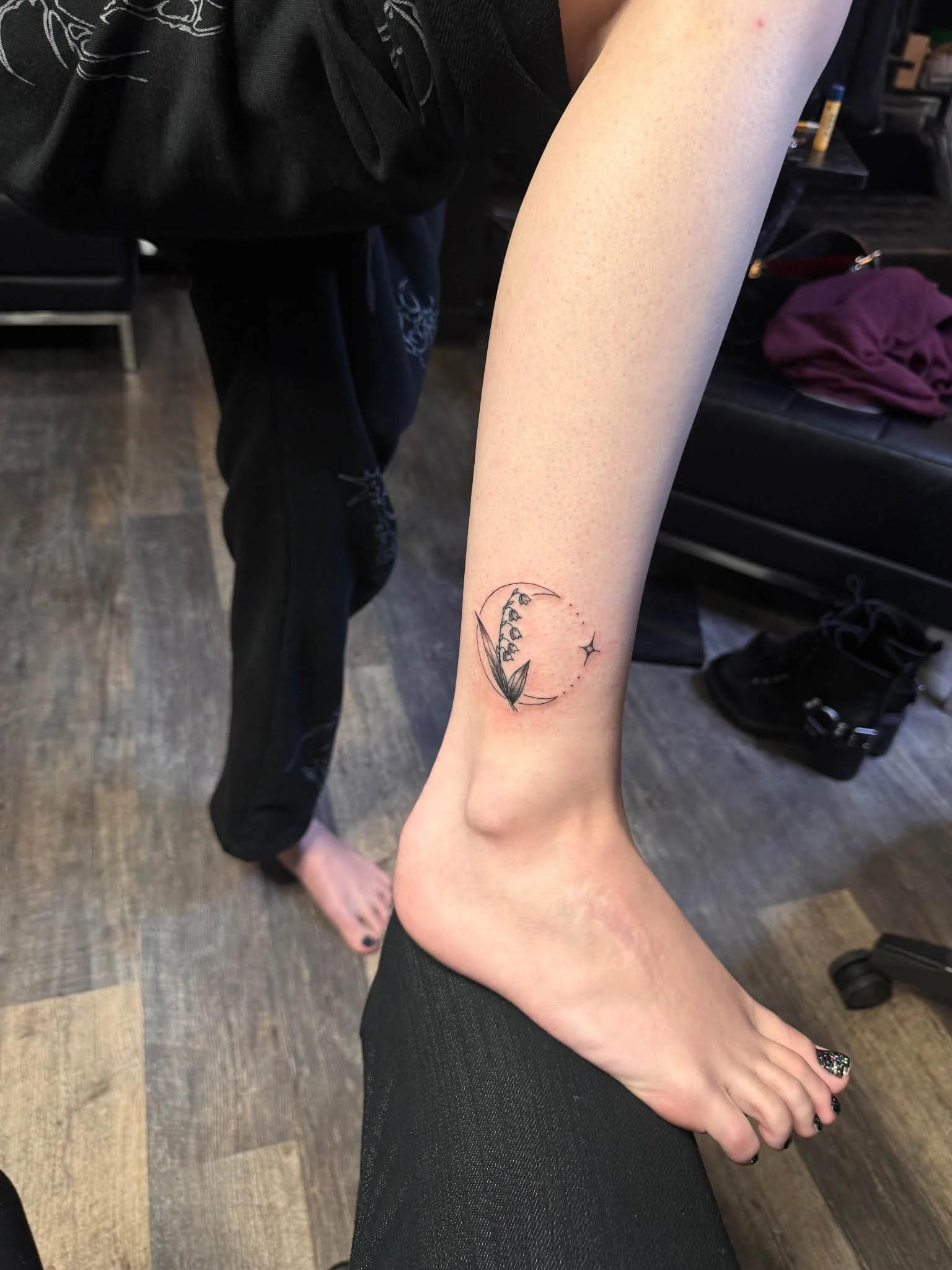 Delicate fine-line tattoo of a lily of the valley flower stem set inside a subtle circular frame with a small starburst accent, located on the lower leg