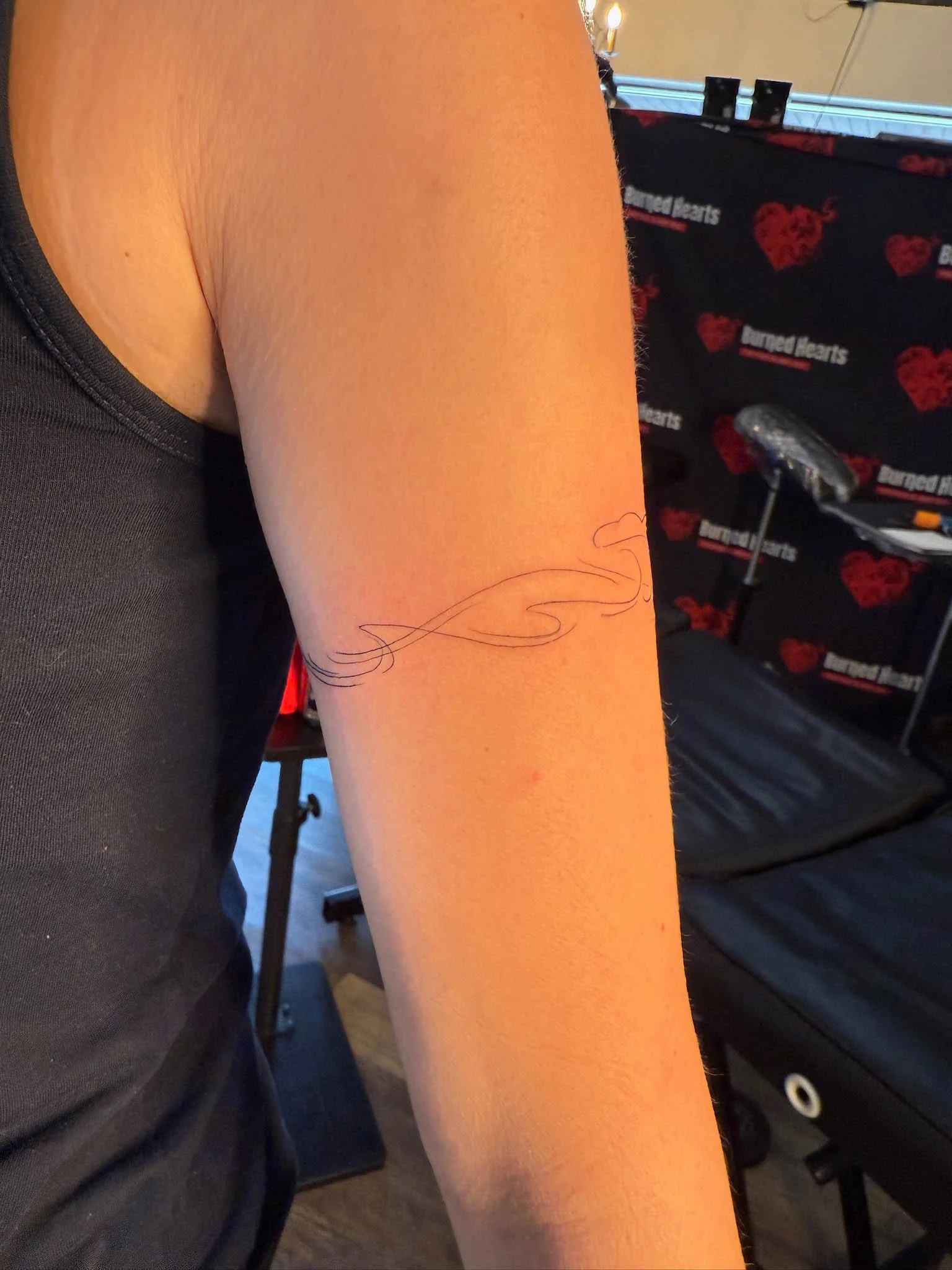 Close-up of a person's arm with a tattoo of a thin, flowing line design in black ink, in an indoor setting with a backdrop that has the 'burned hearts' logo with red hearts and text.