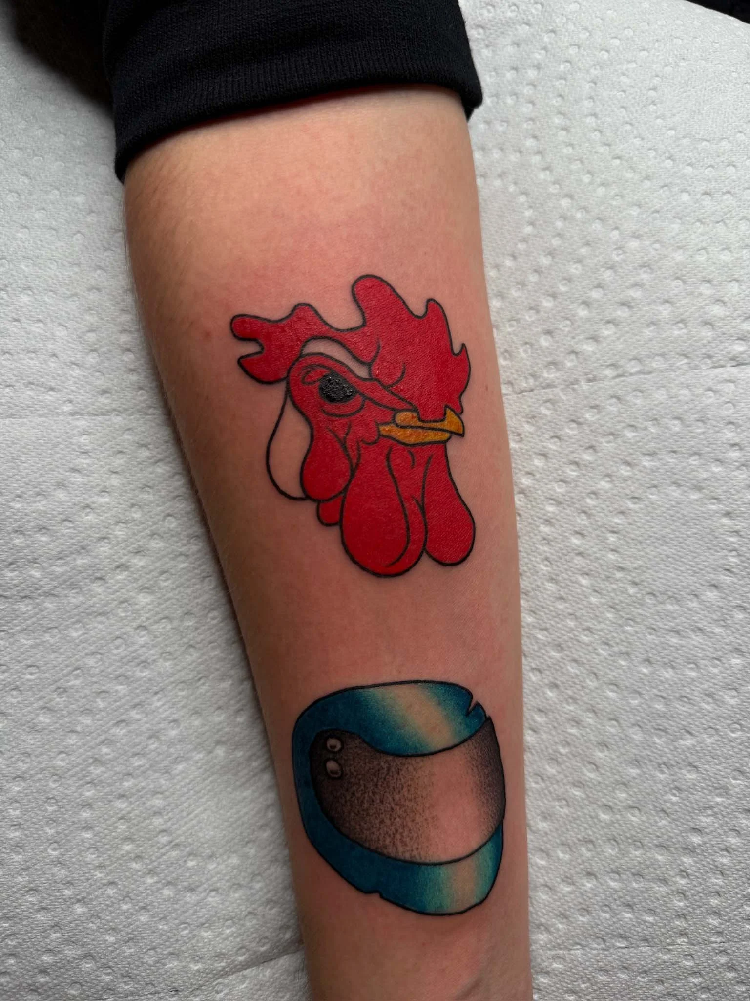 Tattoo of a cartoon rooster and a stylized egg on a person's forearm.