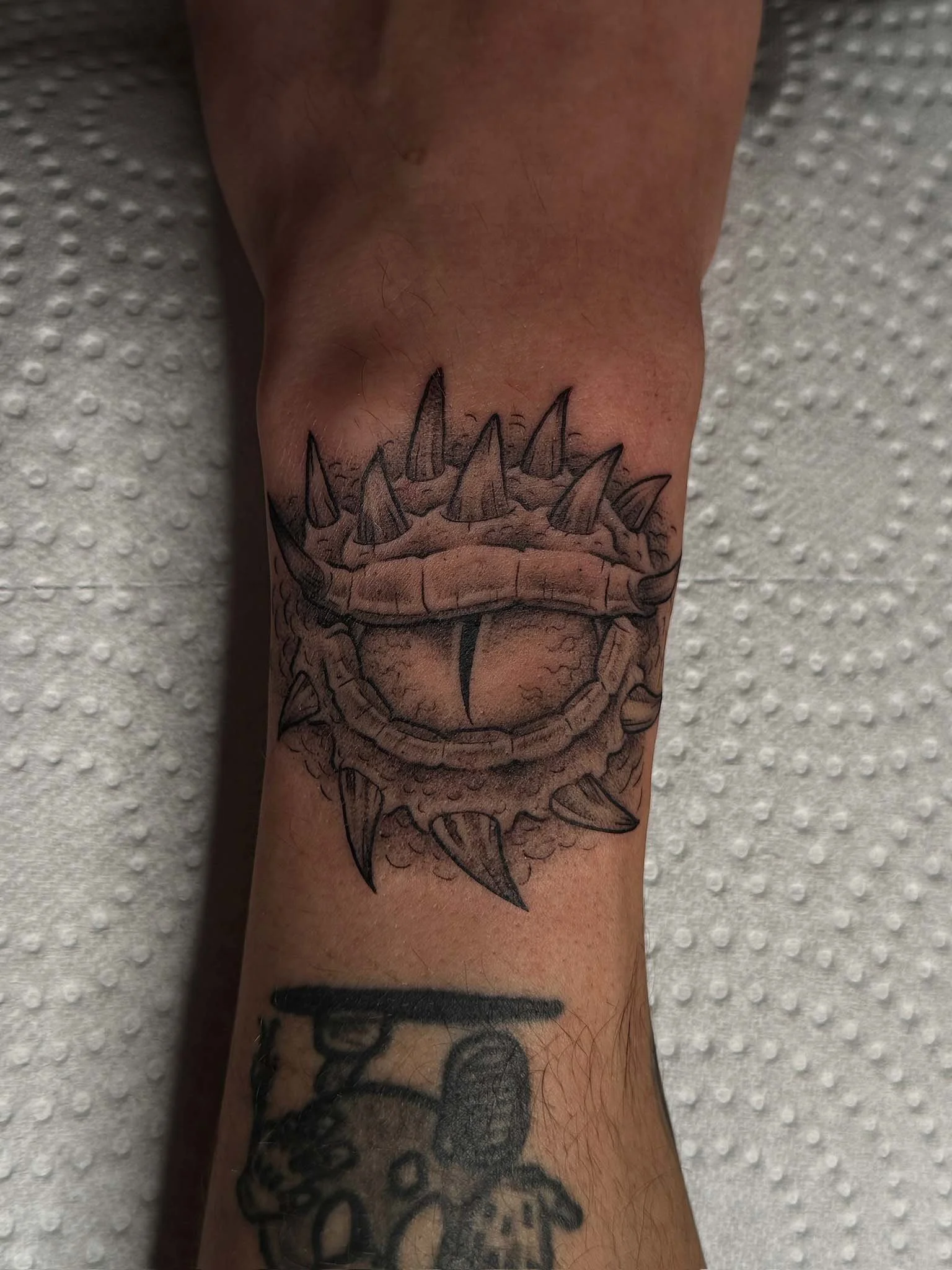 Black and gray tattoo of a monstrous creature's eye with spikes protruding around it, on a person's forearm.