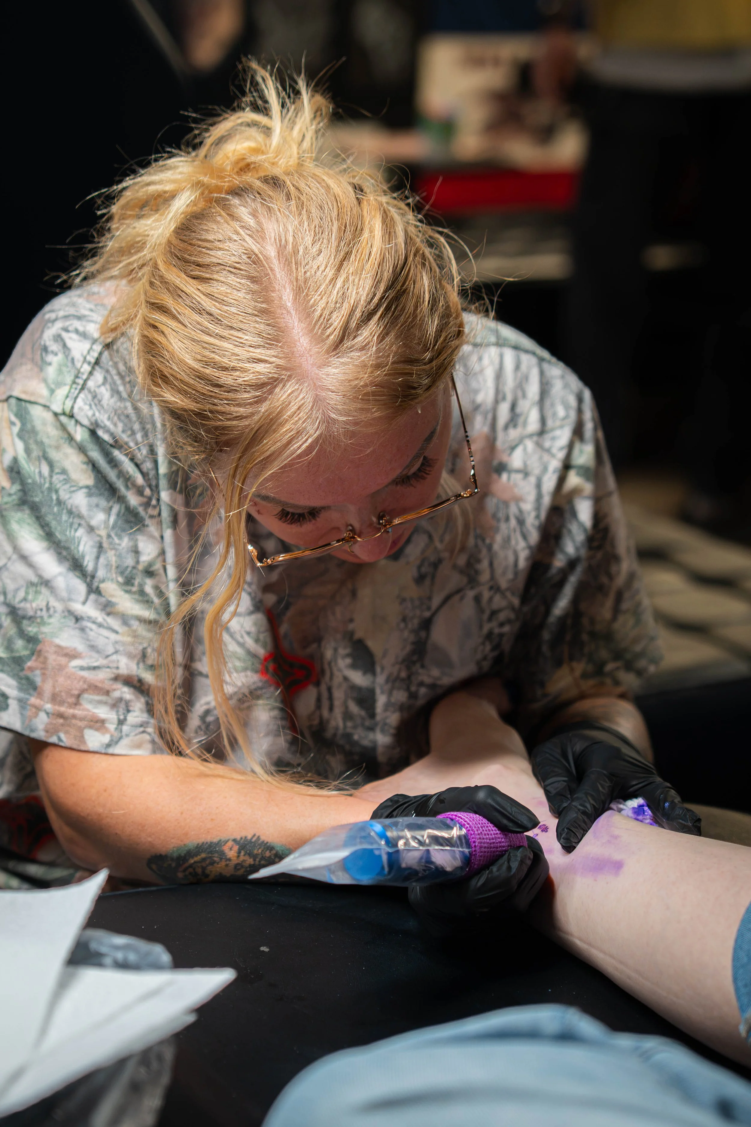 Tattoo artist wearing glasses, using a tattoo machine on a person's arm.