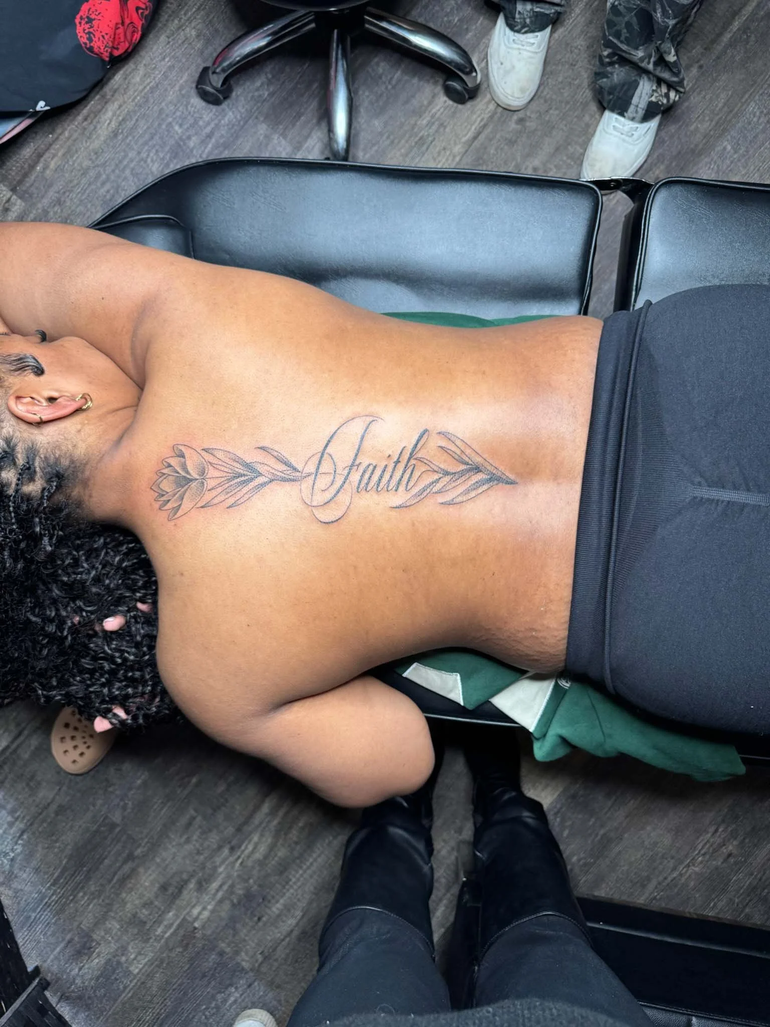 A woman with curly hair and earrings lying face down on a chair in a tattoo studio, showing a large tattoo of the word 'Faith' with decorative flourishes on her upper back and shoulder.
