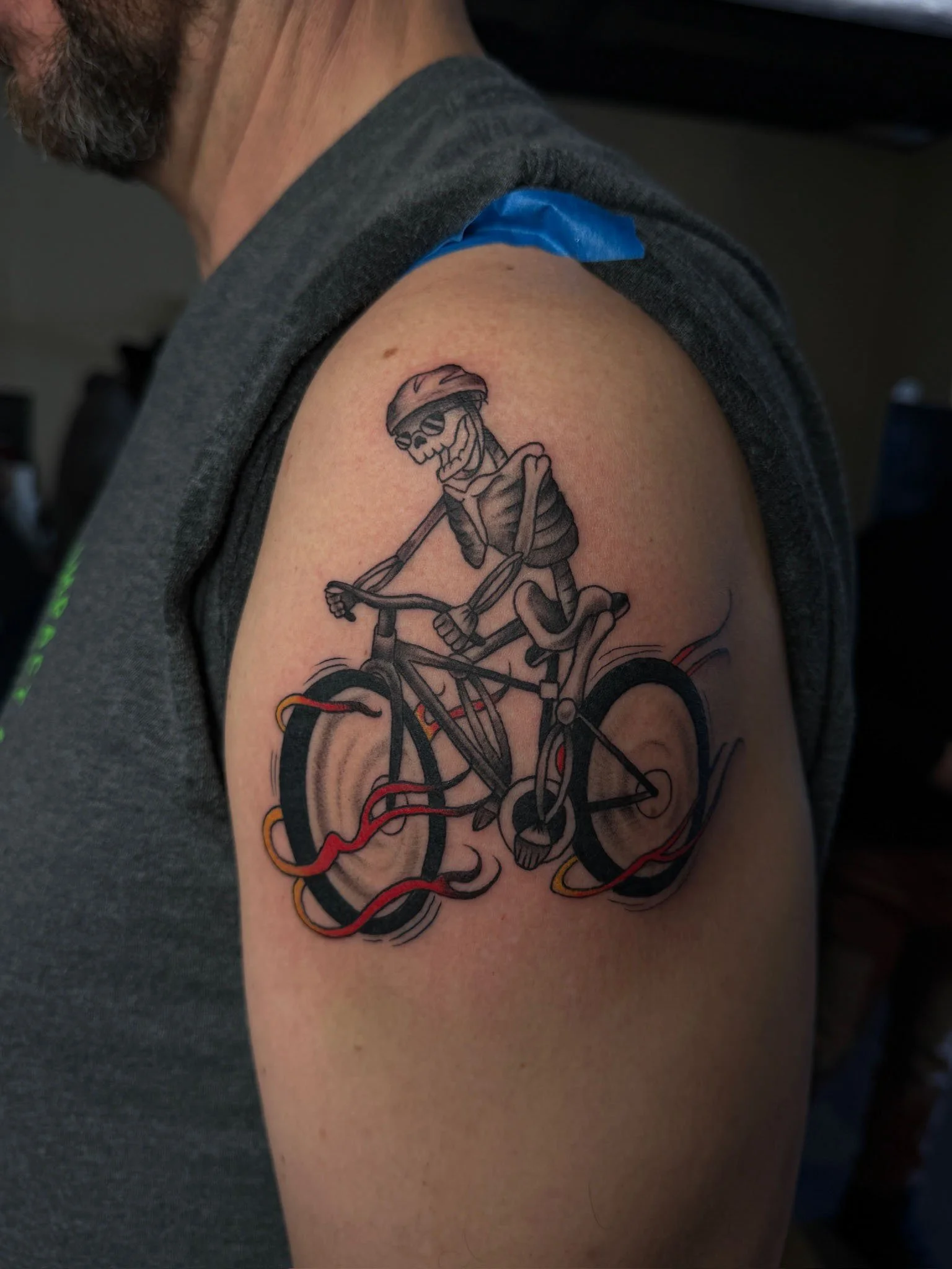 Tattoo of a skeleton riding a bicycle with wires connected to the wheels.