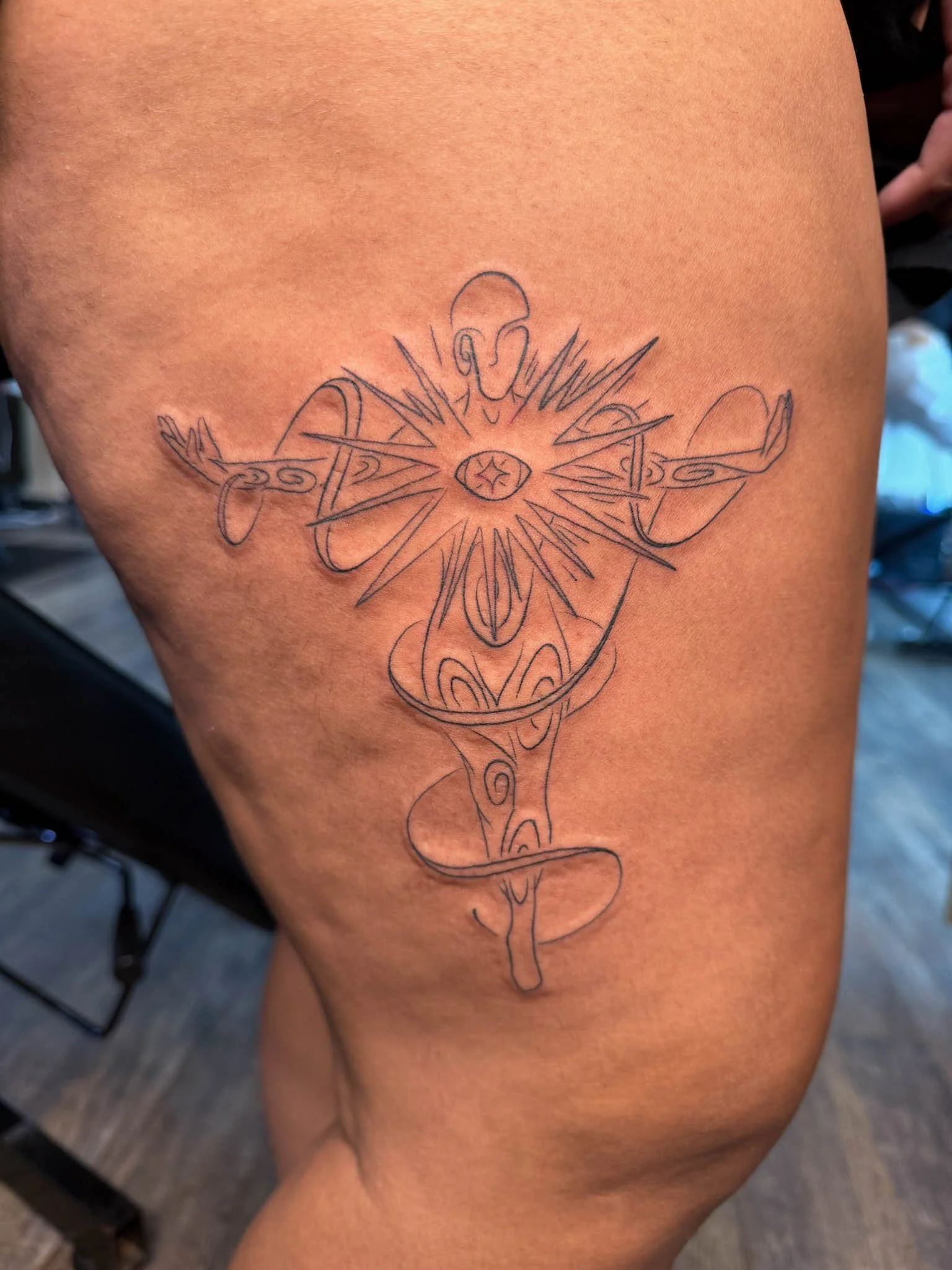Line drawing tattoo of a figure with outstretched arms, an eye on the chest, and a snake wrapping around the body, on a person's thigh.