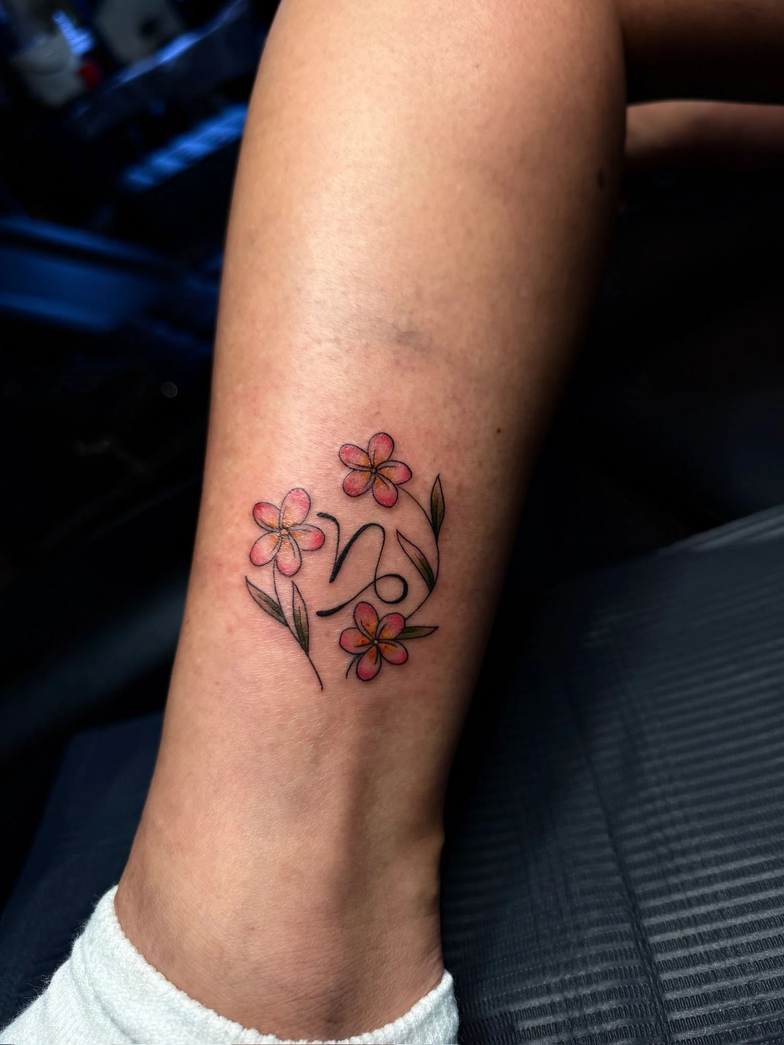 Tattoo of pink flowers and green leaves surrounding a cursive letter N on a person's lower leg.