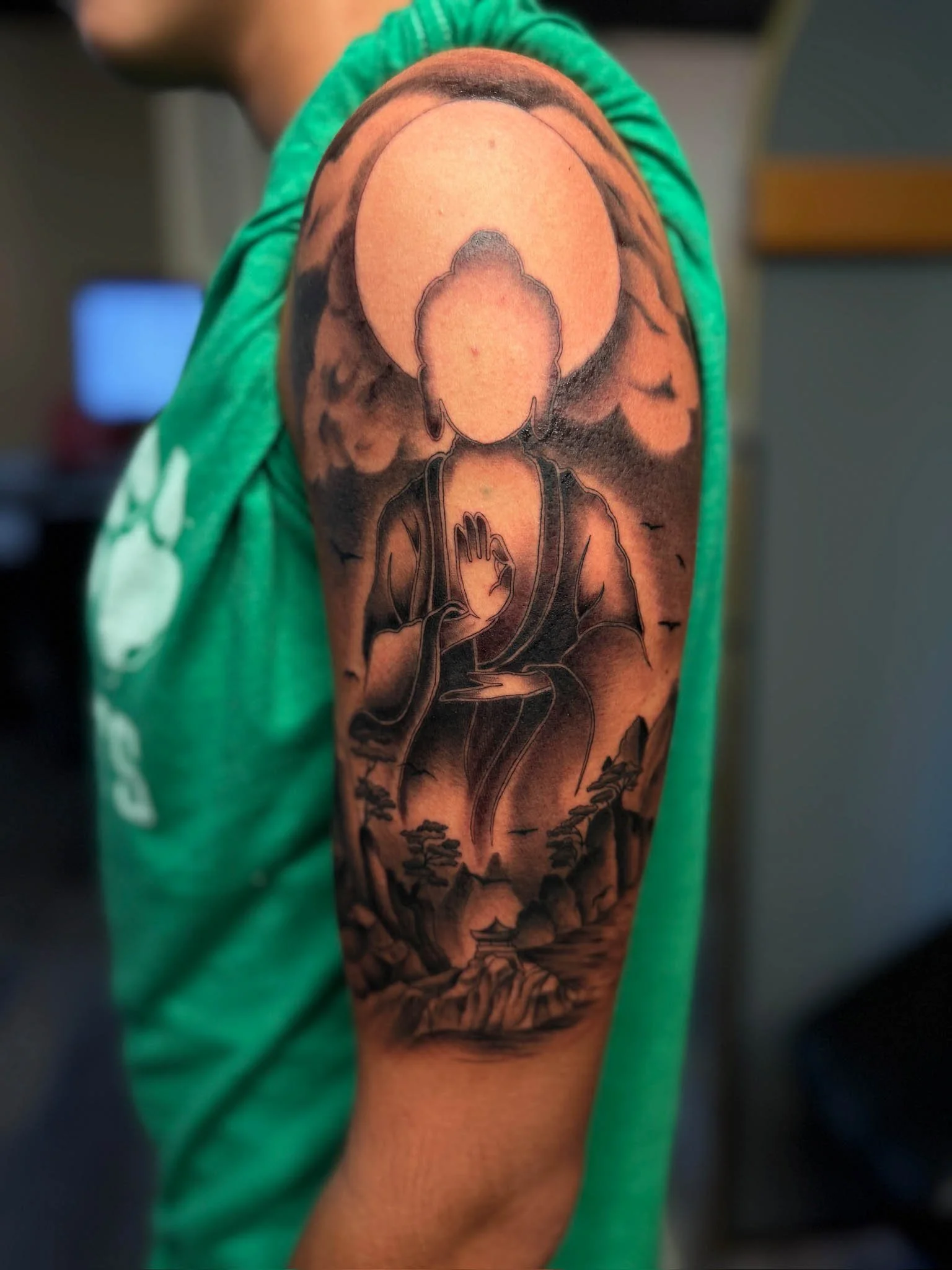 Tattoo of a seated Buddha with a large moon behind it, set against a cloudy sky with mountains, trees, and a pagoda.