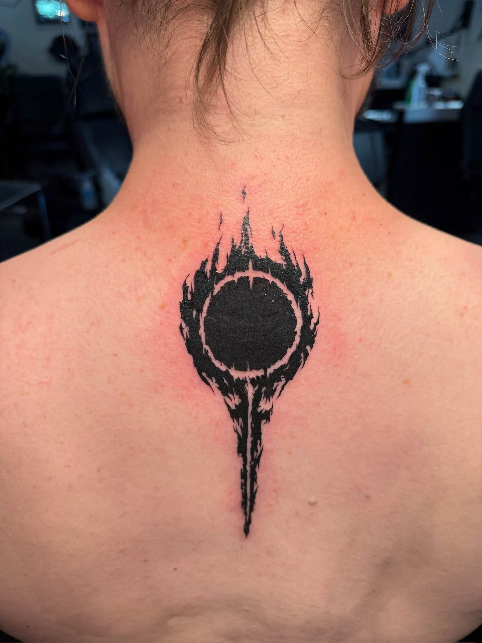Black tattoo on upper back resembling a fiery eye with a central dark circle surrounded by flames and a downward point.