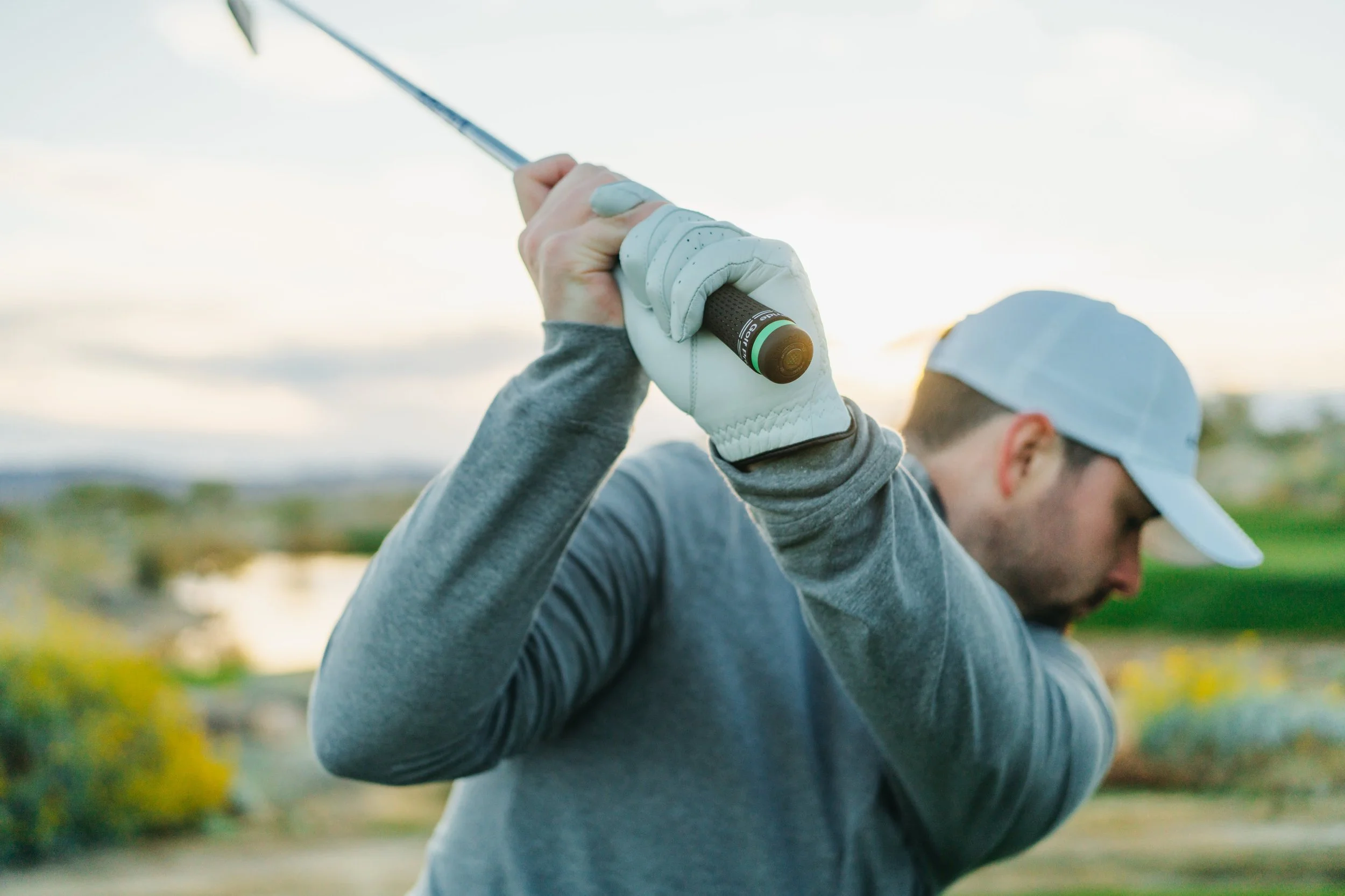 A man swinging a golf club outdoors during sunset, wearing a white cap and sportswear.