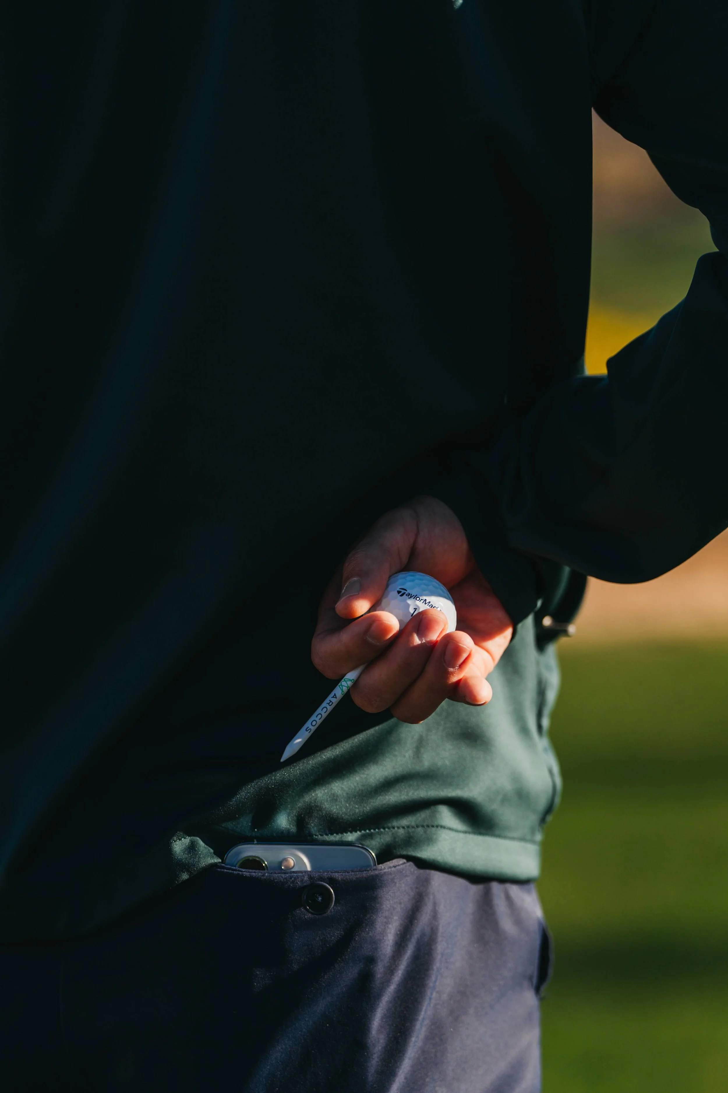 A person holds a golf ball and tee, with a cell phone partially visible in their pocket, dressed in dark clothing and pants, standing outdoors.