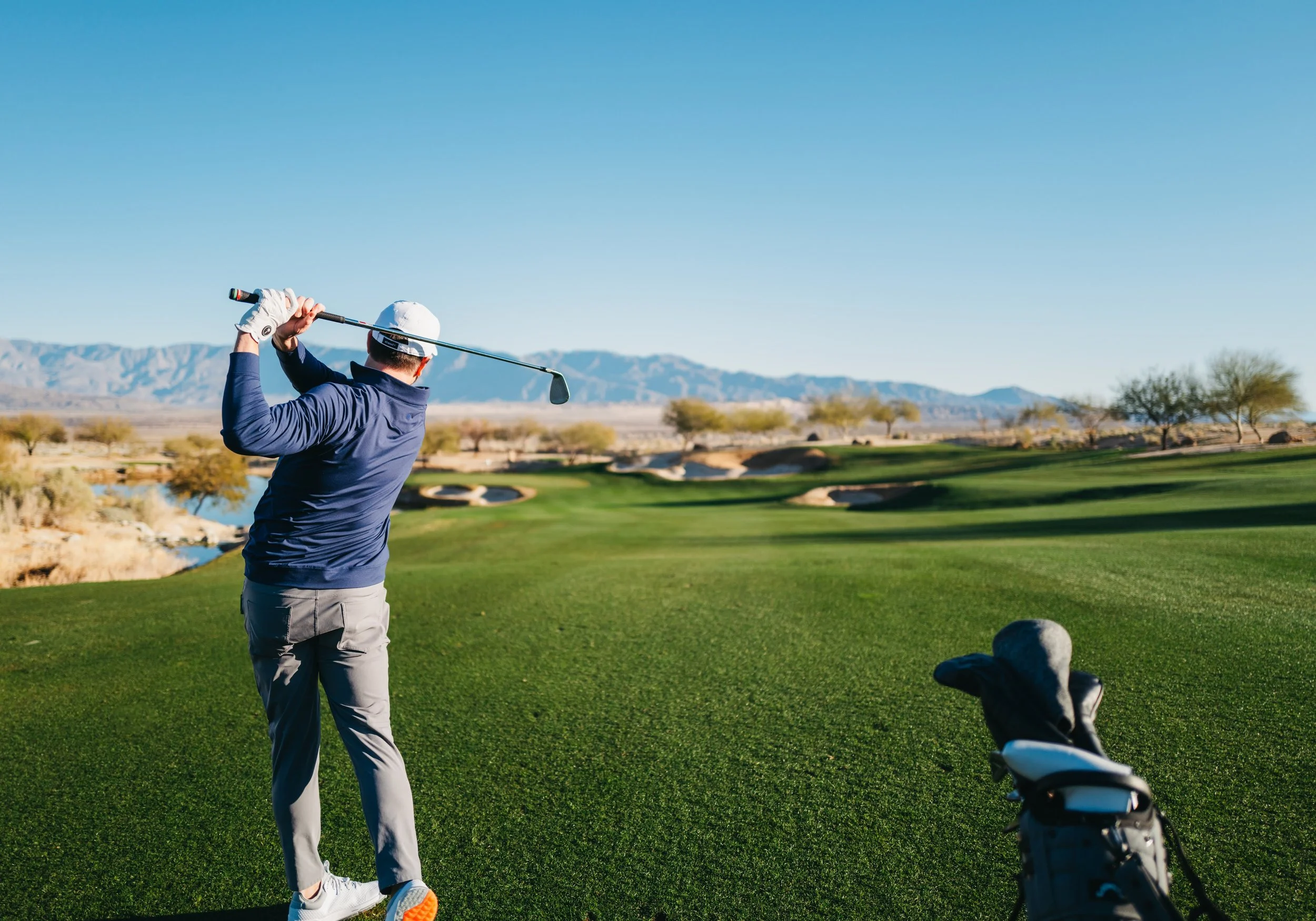 Man in navy jacket and gray pants swinging a golf club on a golf course with a golf bag nearby, desert landscape with mountains in the background.