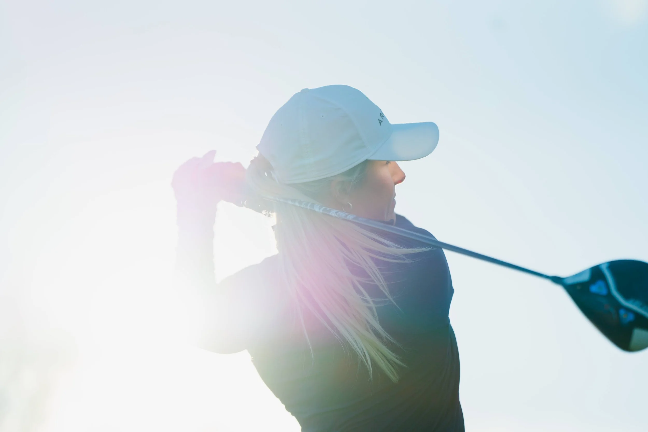 A woman playing golf swings a club on a golf course, wearing a cap and sunglasses, with bright sunlight in the background.