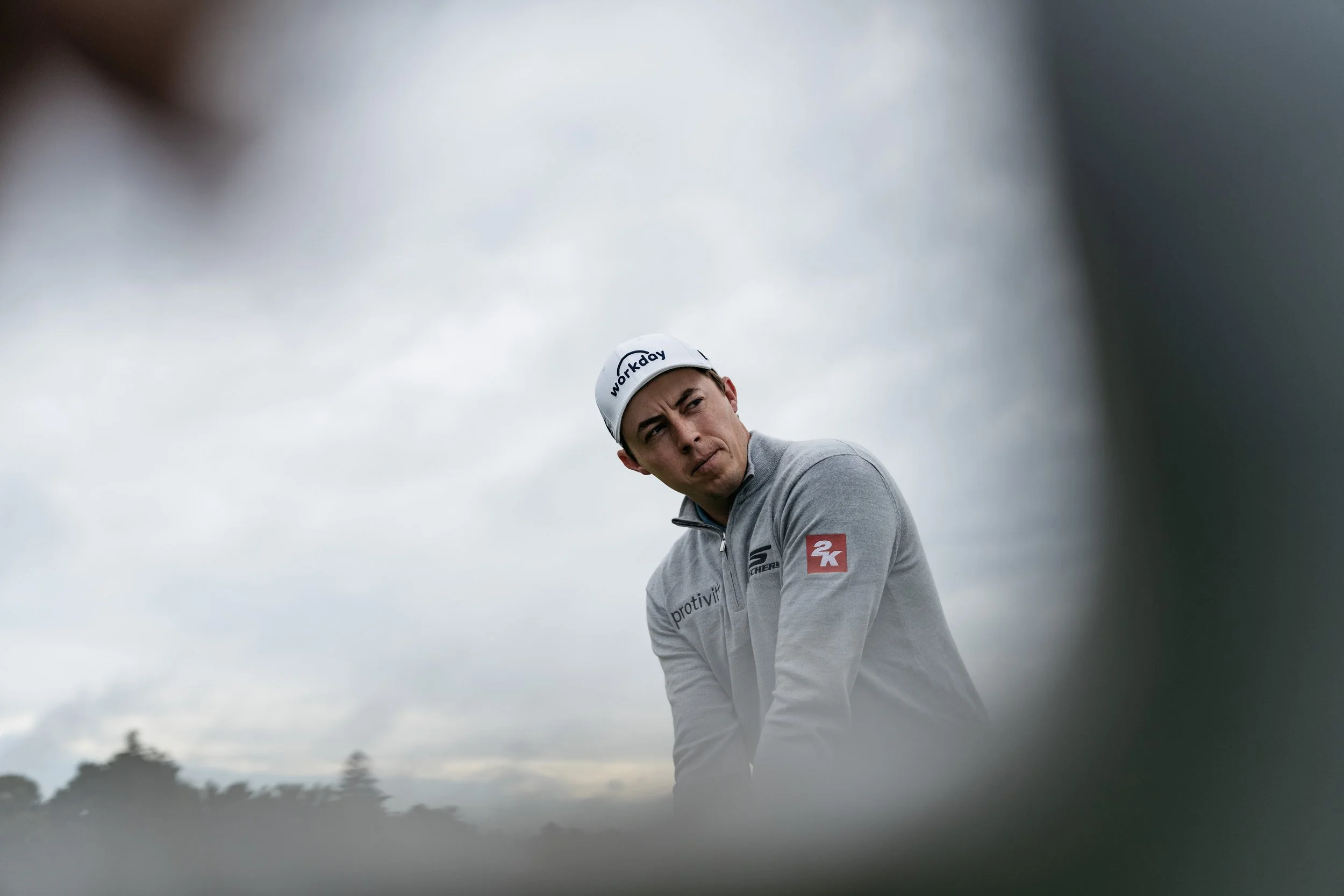 A male golfer in a cap and grey jacket looking to the side, shot from a low angle with blurred foreground elements and cloudy sky background.