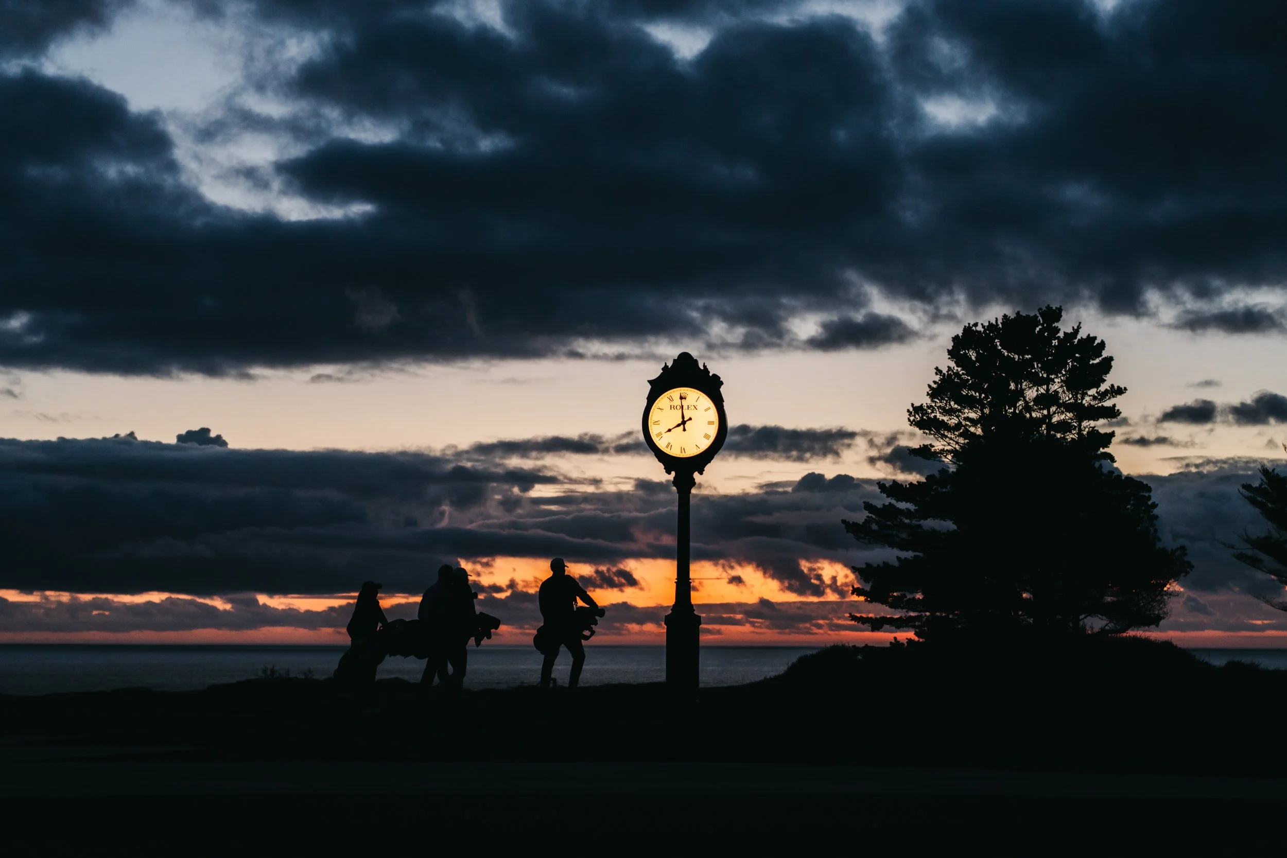 Silhouettes of three people and a horse standing near a clock tower during sunset, with dark clouds overhead and a tree on the right.