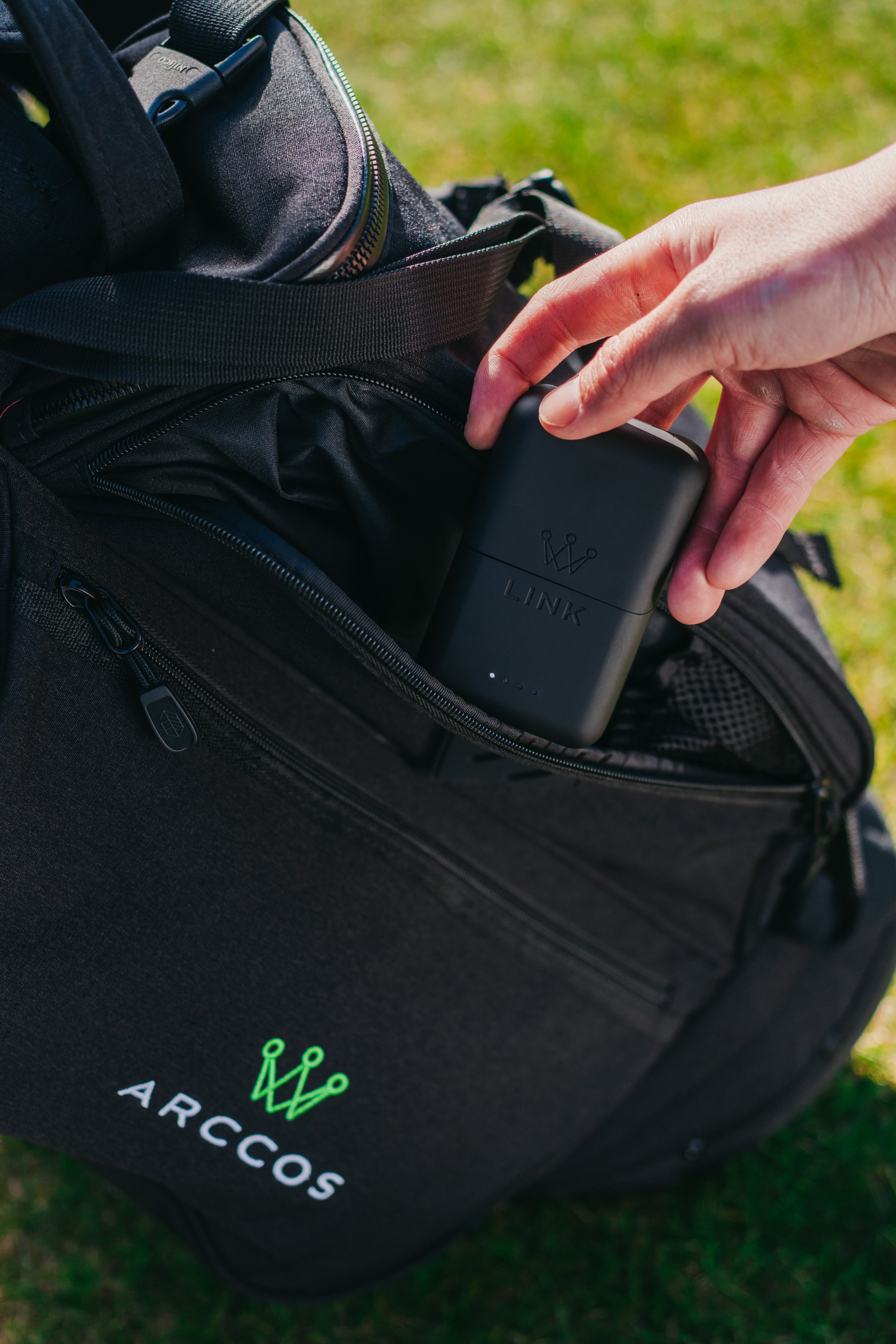 A person placing a portable black power bank with a crown logo and the word 'LINK' into the front pocket of a black ARCCOS branded backpack.