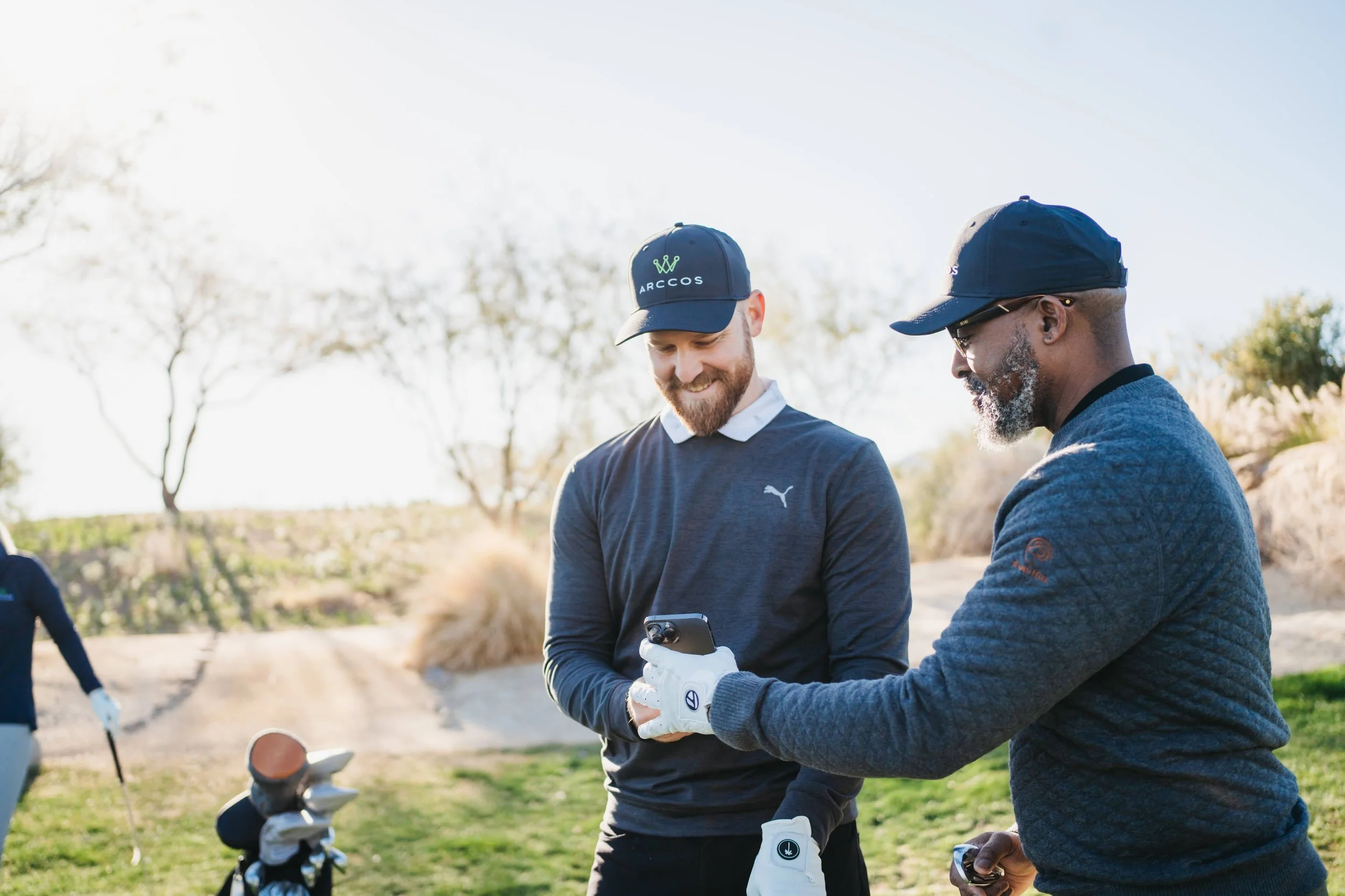 Two men on a golf course looking at a phone together, one with a beard, wearing a black Puma shirt and cap, the other with glasses, wearing a dark baseball cap and gray sweater.