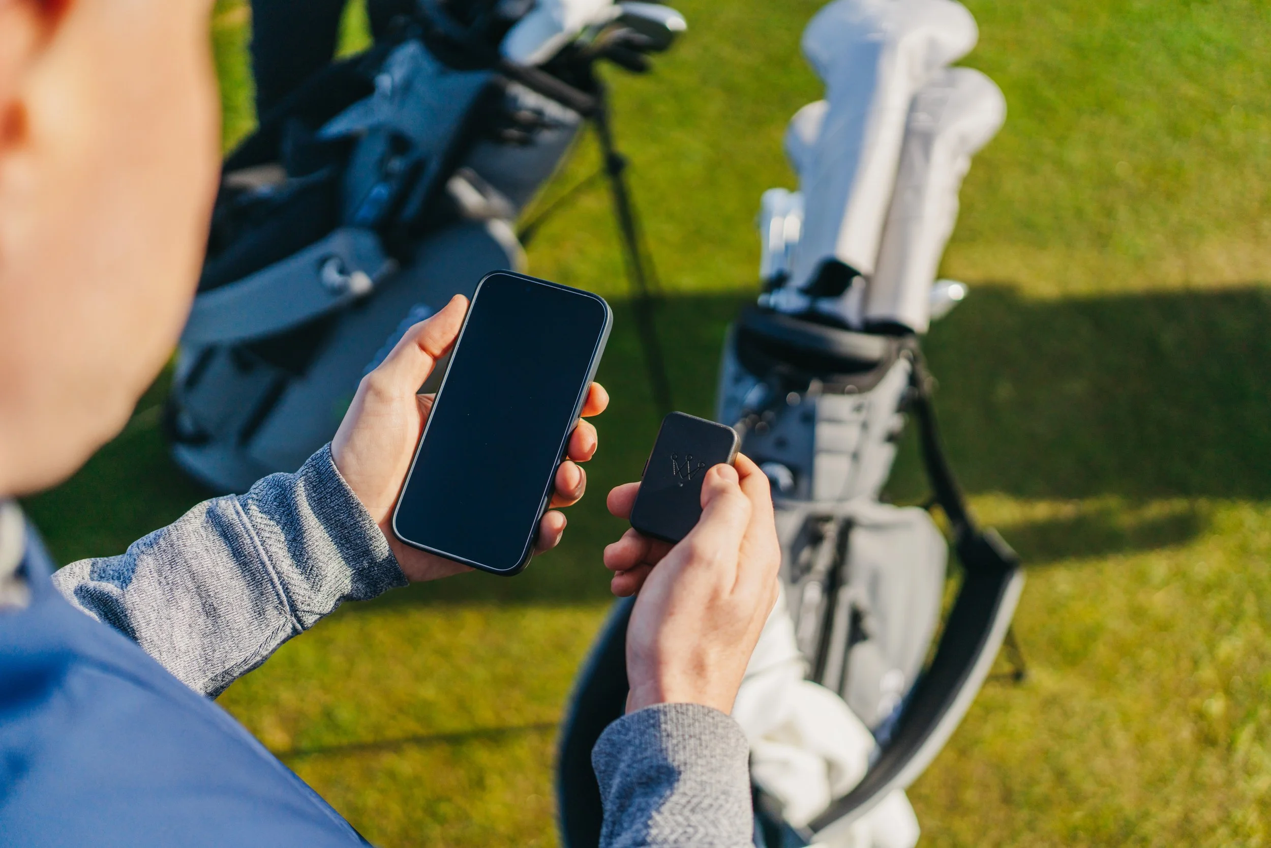 A person holding a smartphone and a portable device near a golf bag with clubs on a golf course.