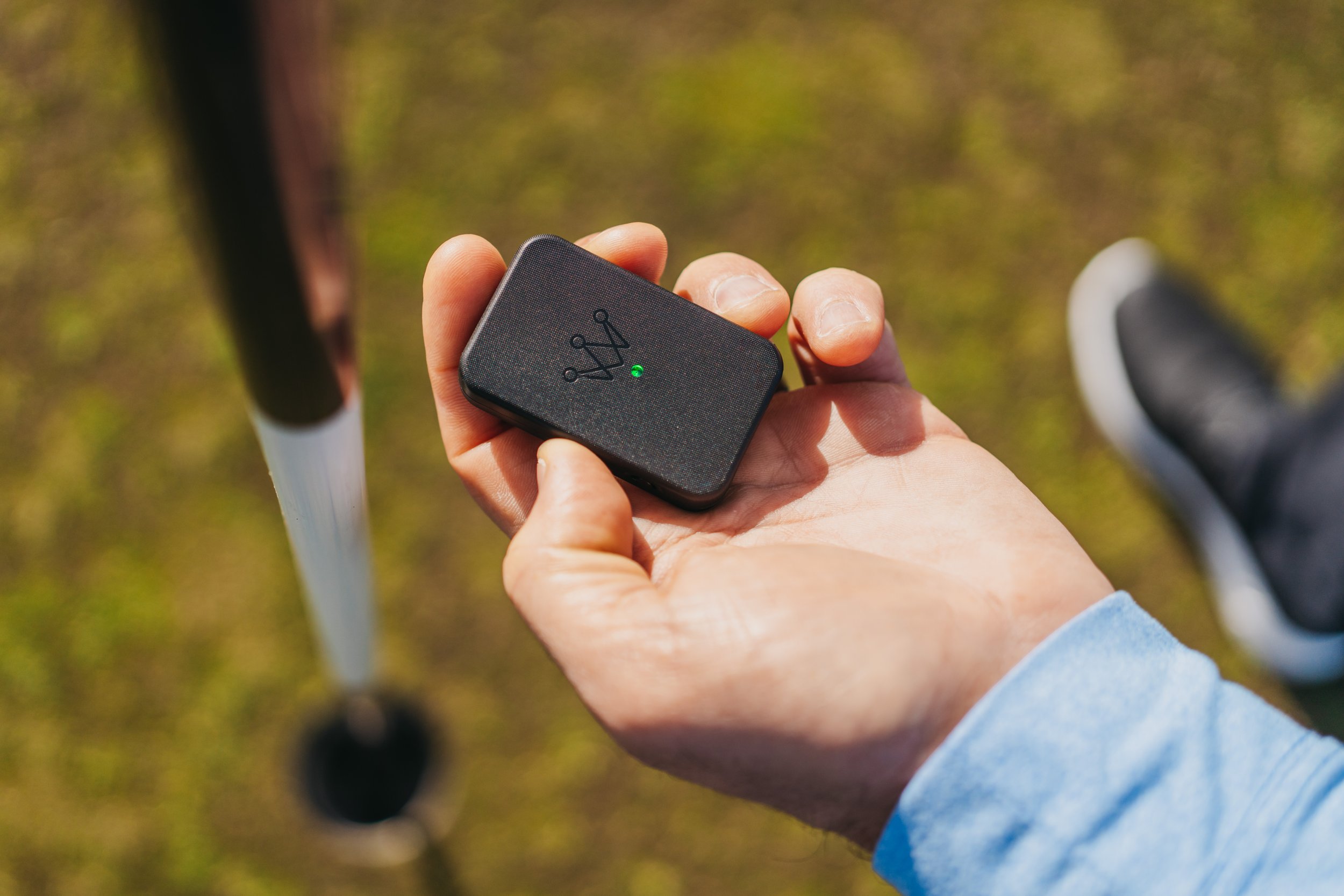 A person holding a small black device with a green indicator light, outdoors on a grassy area with a walker and a shoe visible in the background.