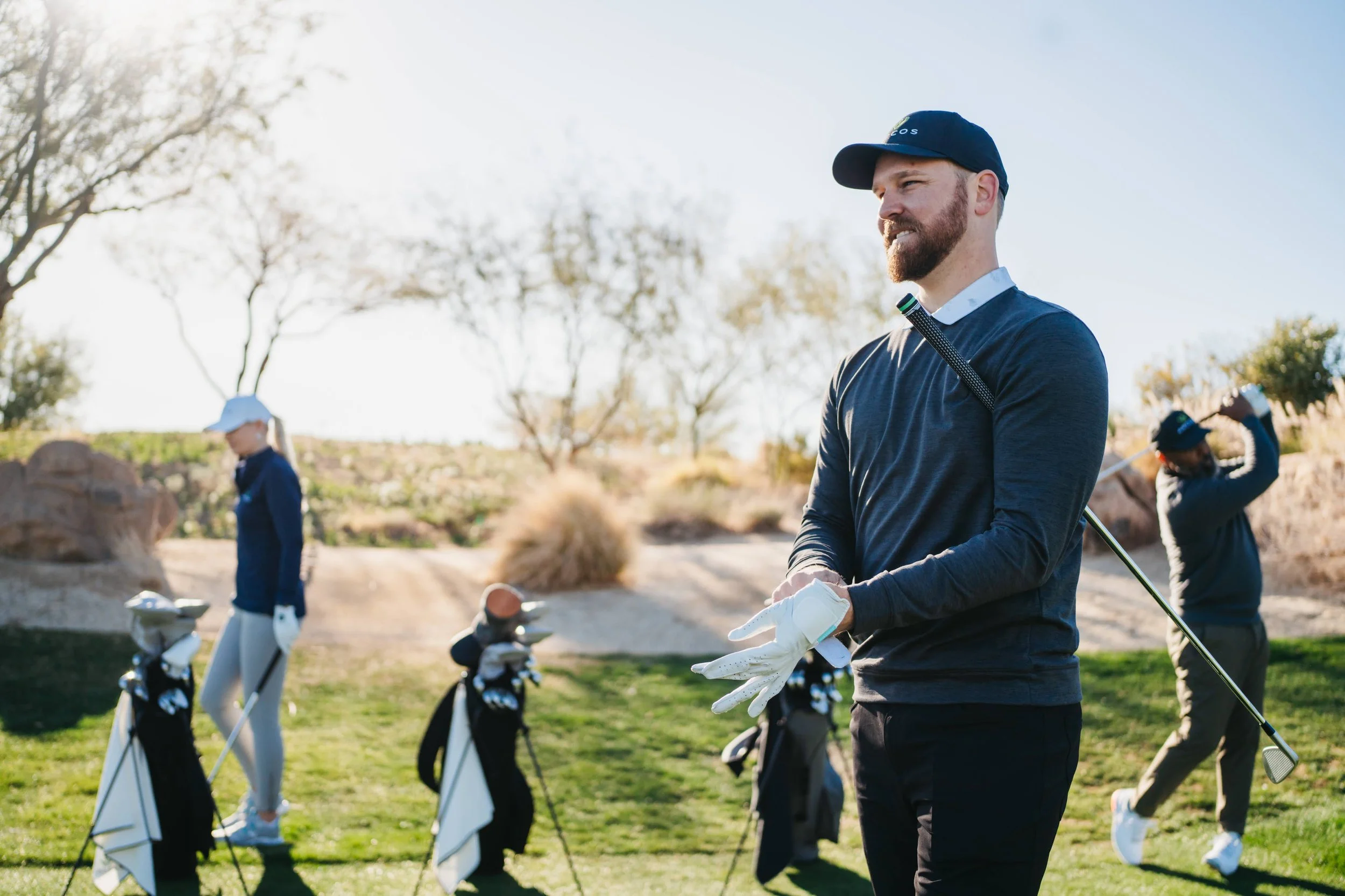 A man in a dark long-sleeve shirt, black cap, and white golf glove standing on a golf course, holding a golf club, with two other people practicing swings in the background, trees and rocks visible.