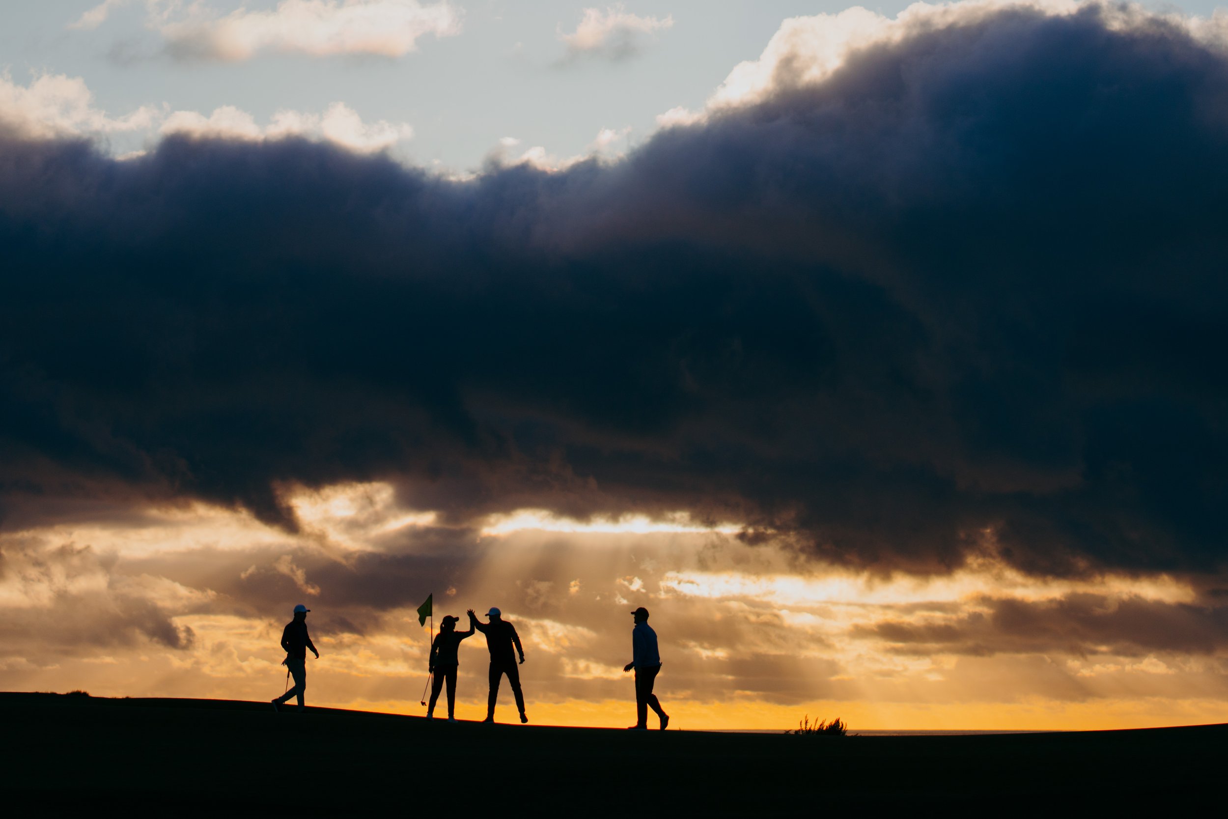 Silhouettes of four people on a golf course at sunset, with dark clouds overhead and rays of sunlight breaking through.