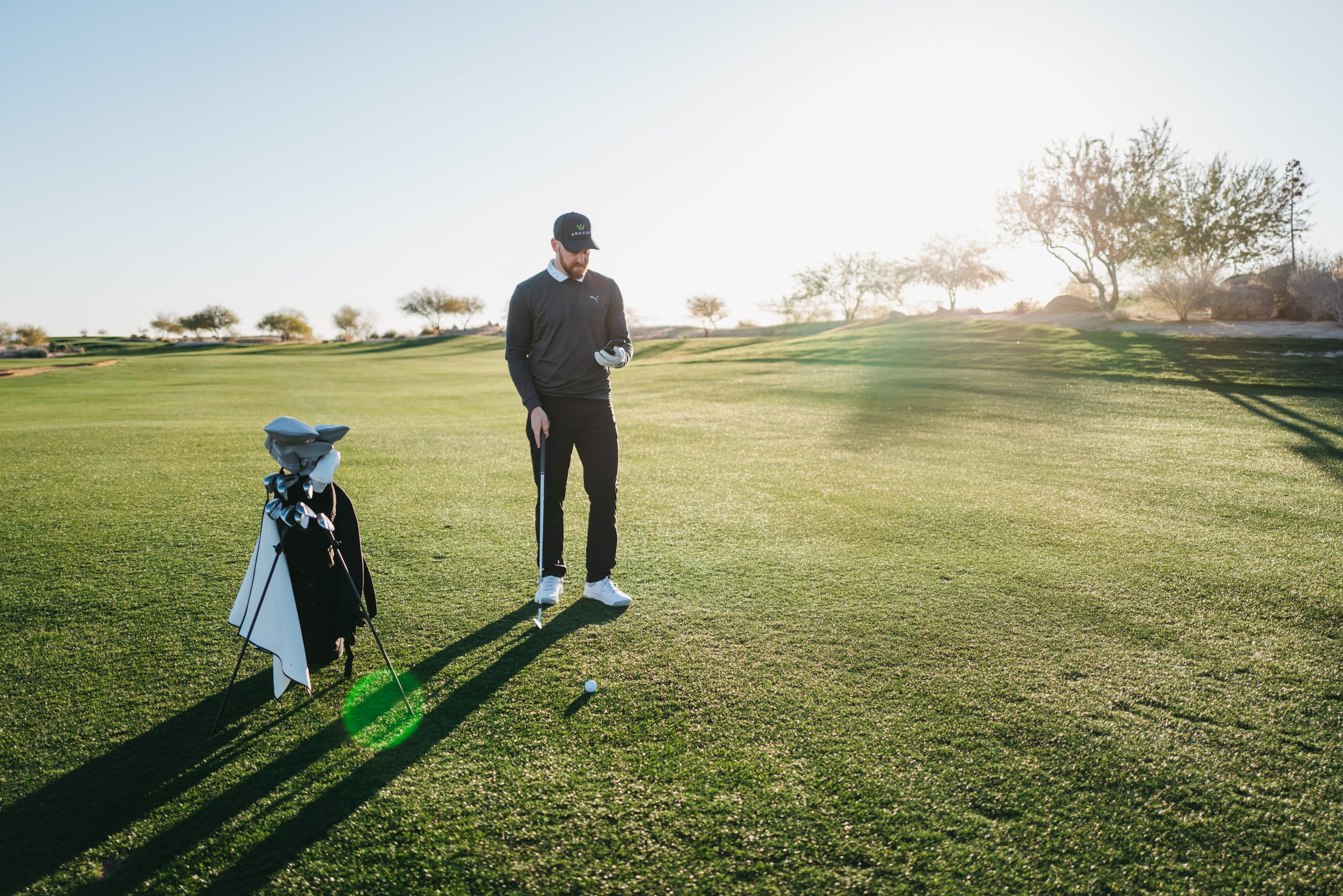 A golfer stands on a golf course preparing to hit a golf ball, with a golf bag placed on a stand nearby, illuminated by bright sunlight.