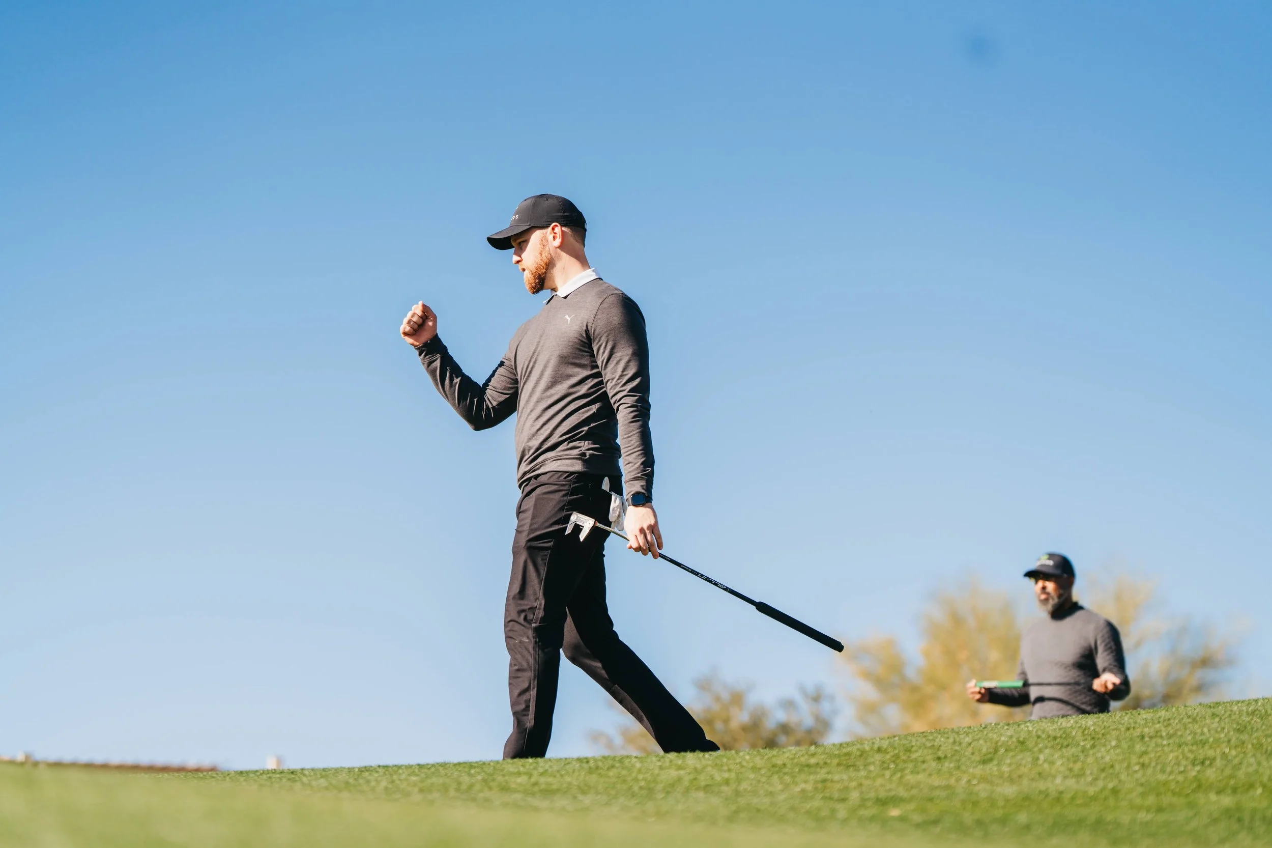 A man with a beard wearing a baseball cap, sunglasses, gray long-sleeve shirt, and black pants is walking on the grass holding a golf club, with another man in the background also holding a golf club on a golf course under a clear blue sky.