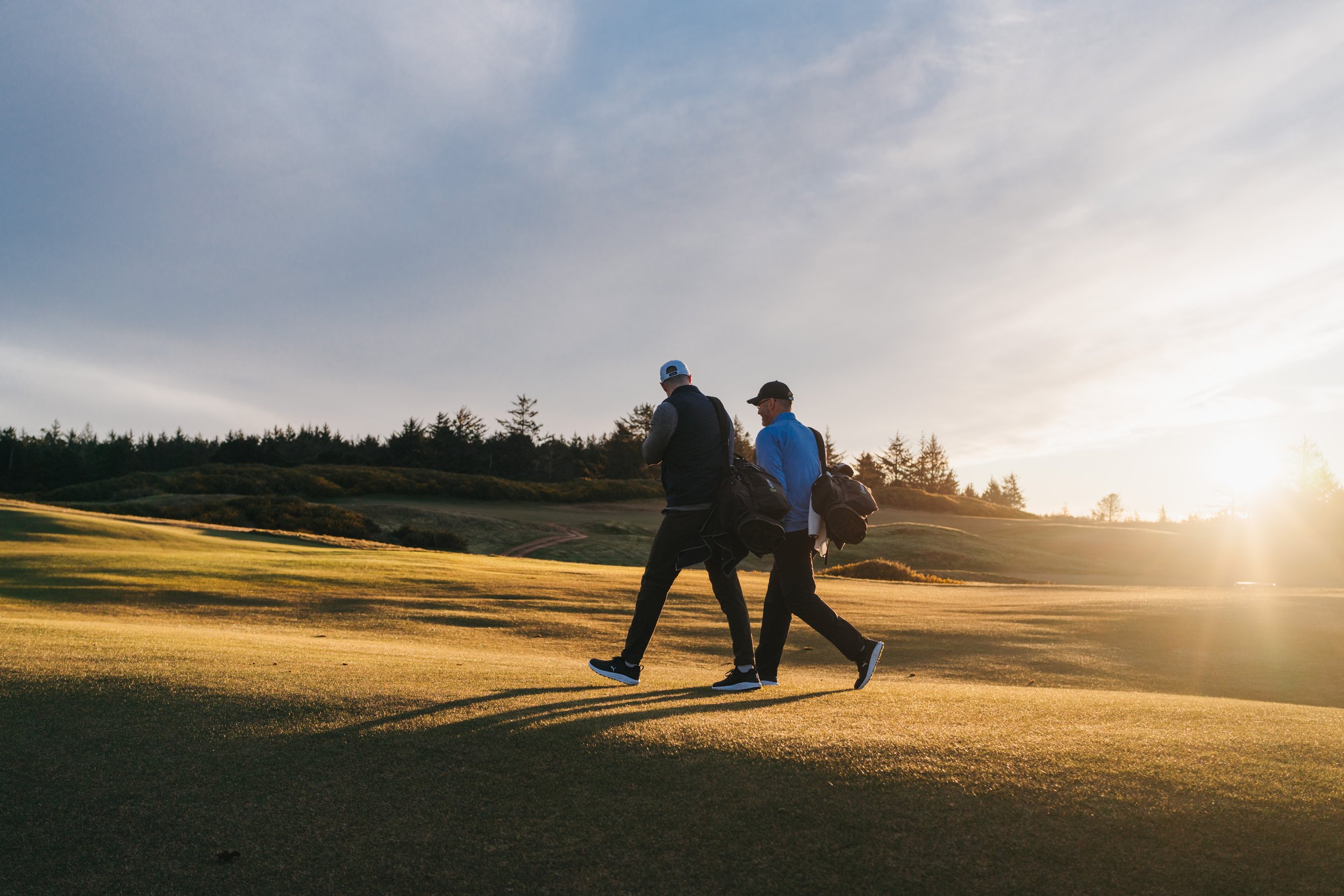 Two men with golf bags walking on a golf course at sunset.