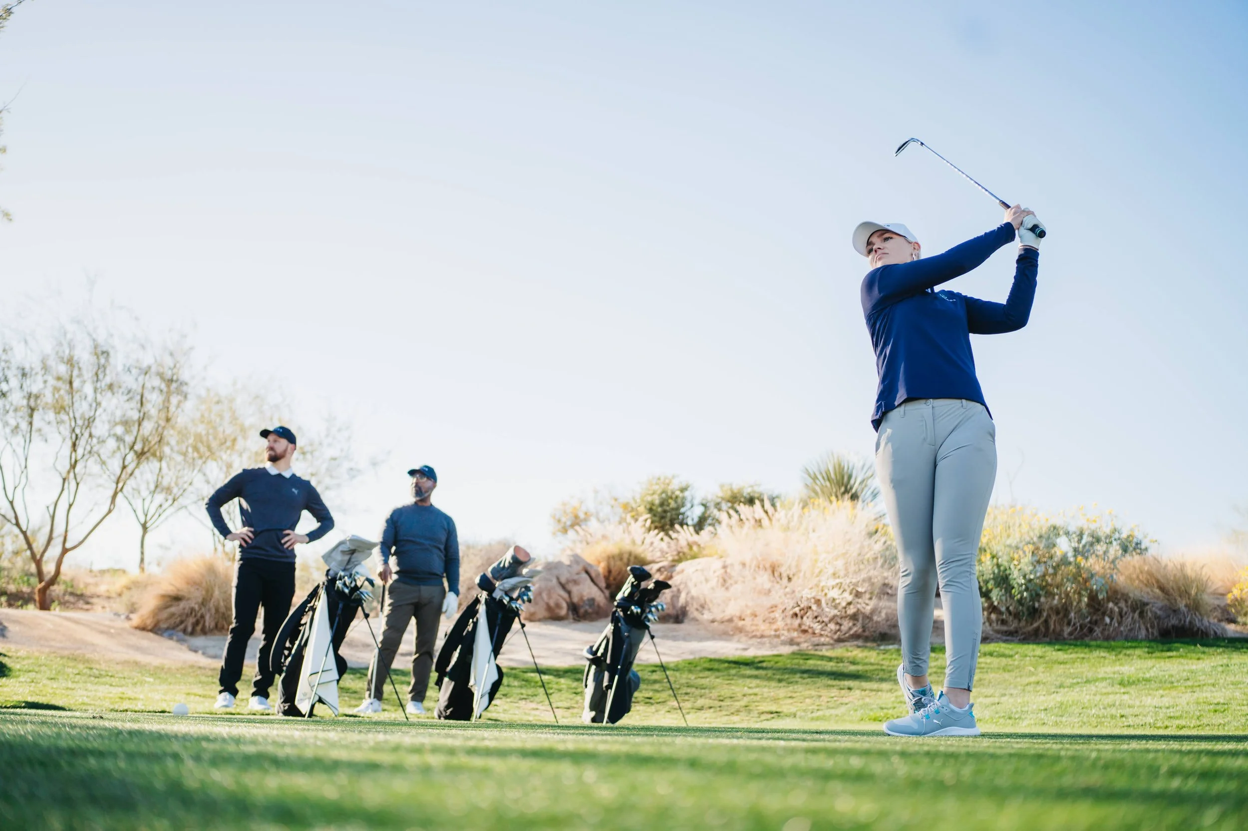 A woman swings a golf club on a golf course while two men watch in the background.