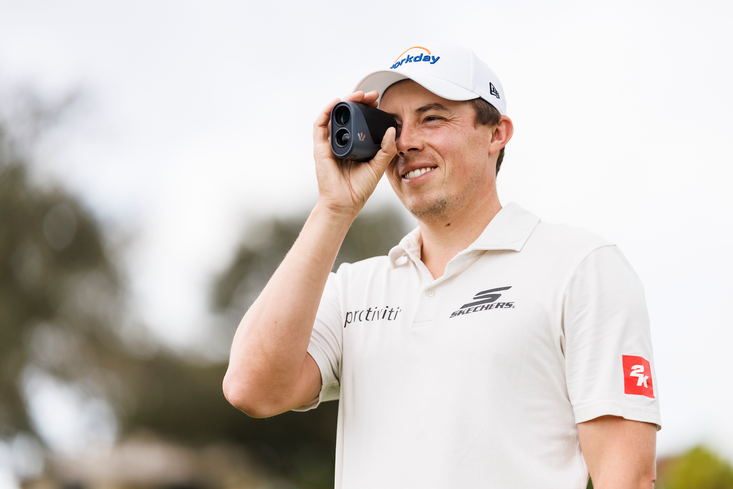 A man in a white polo shirt and white cap using a golf rangefinder outdoors, with trees and a cloudy sky in the background.