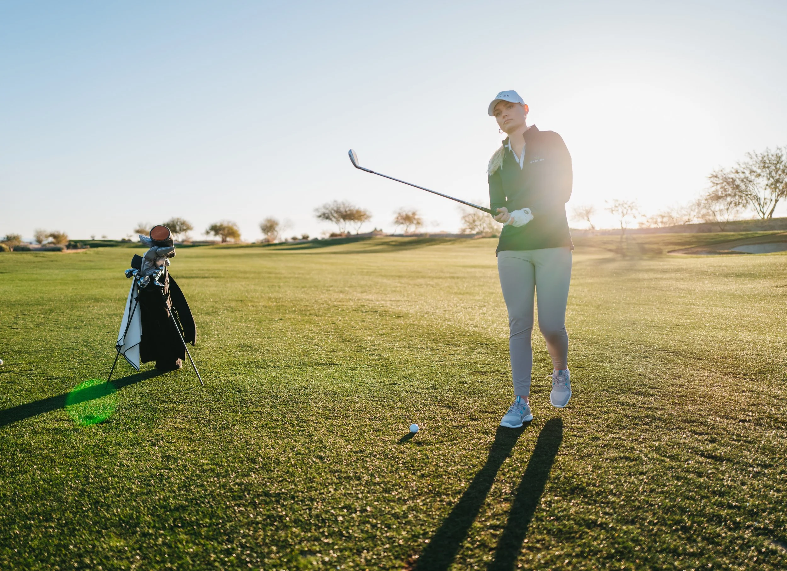 A woman standing on a golf course holding a golf club with a golf bag on the ground nearby, as the sun sets behind her.