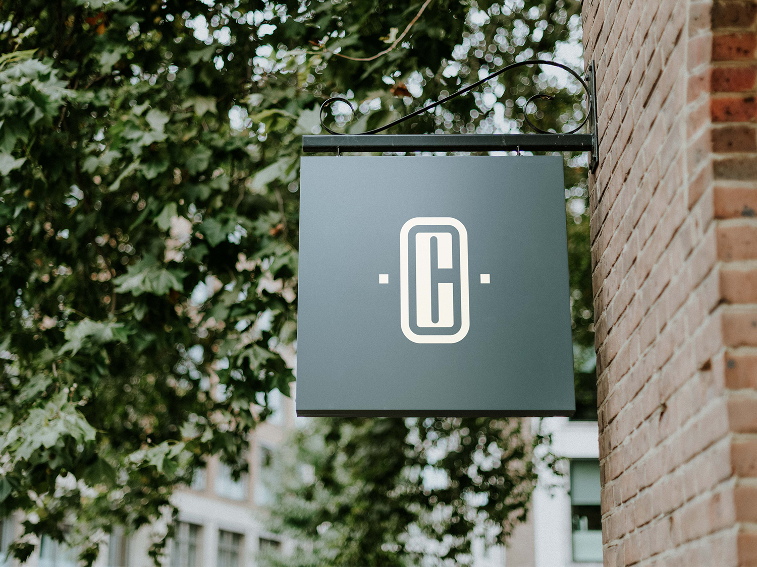 A black and white sign hanging from a bracket on a brick wall, featuring a logo with a stylized letter 'C' inside an oval shape.