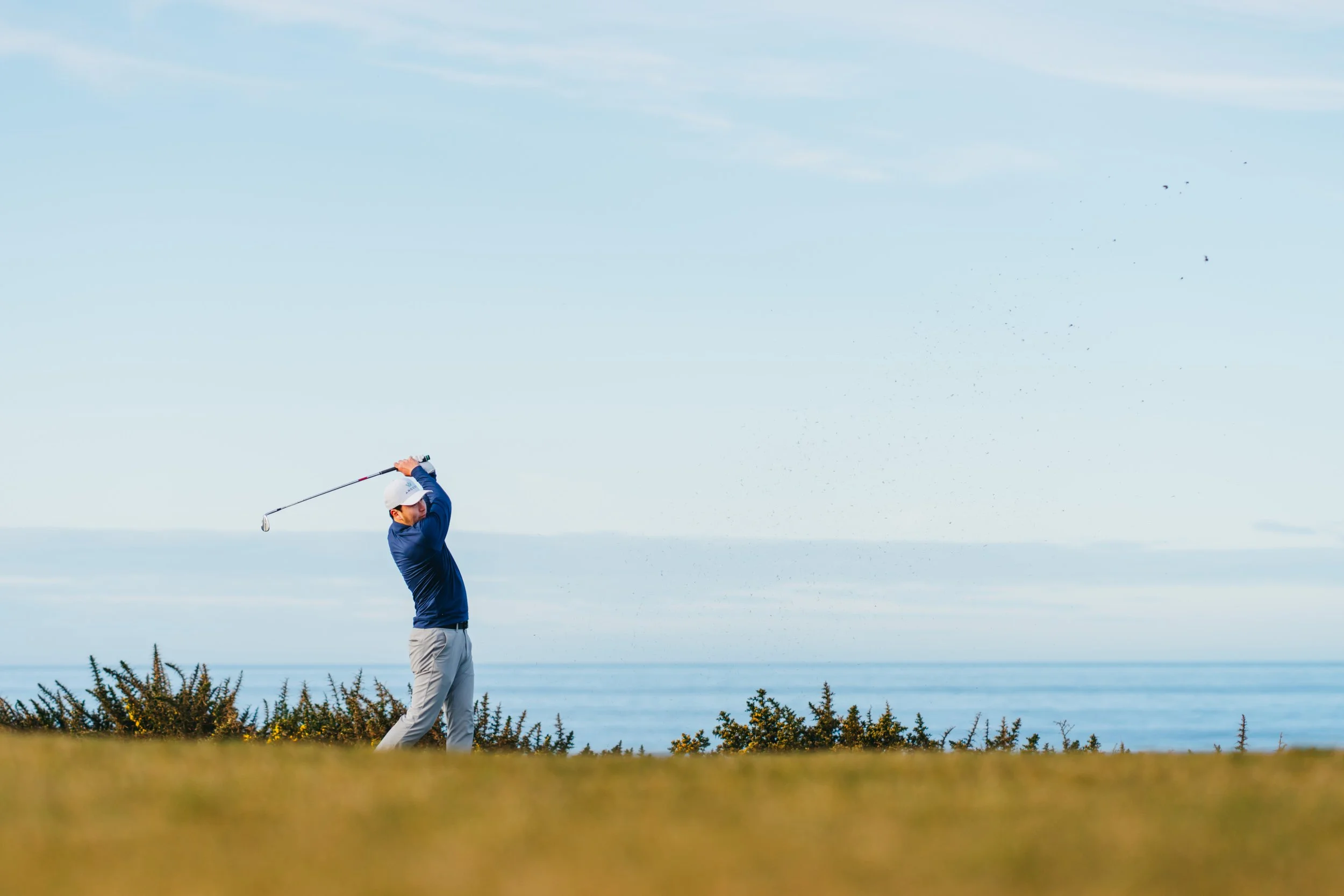 A person playing golf on a course near the coast, swinging a golf club, with a clear blue sky and ocean in the background.
