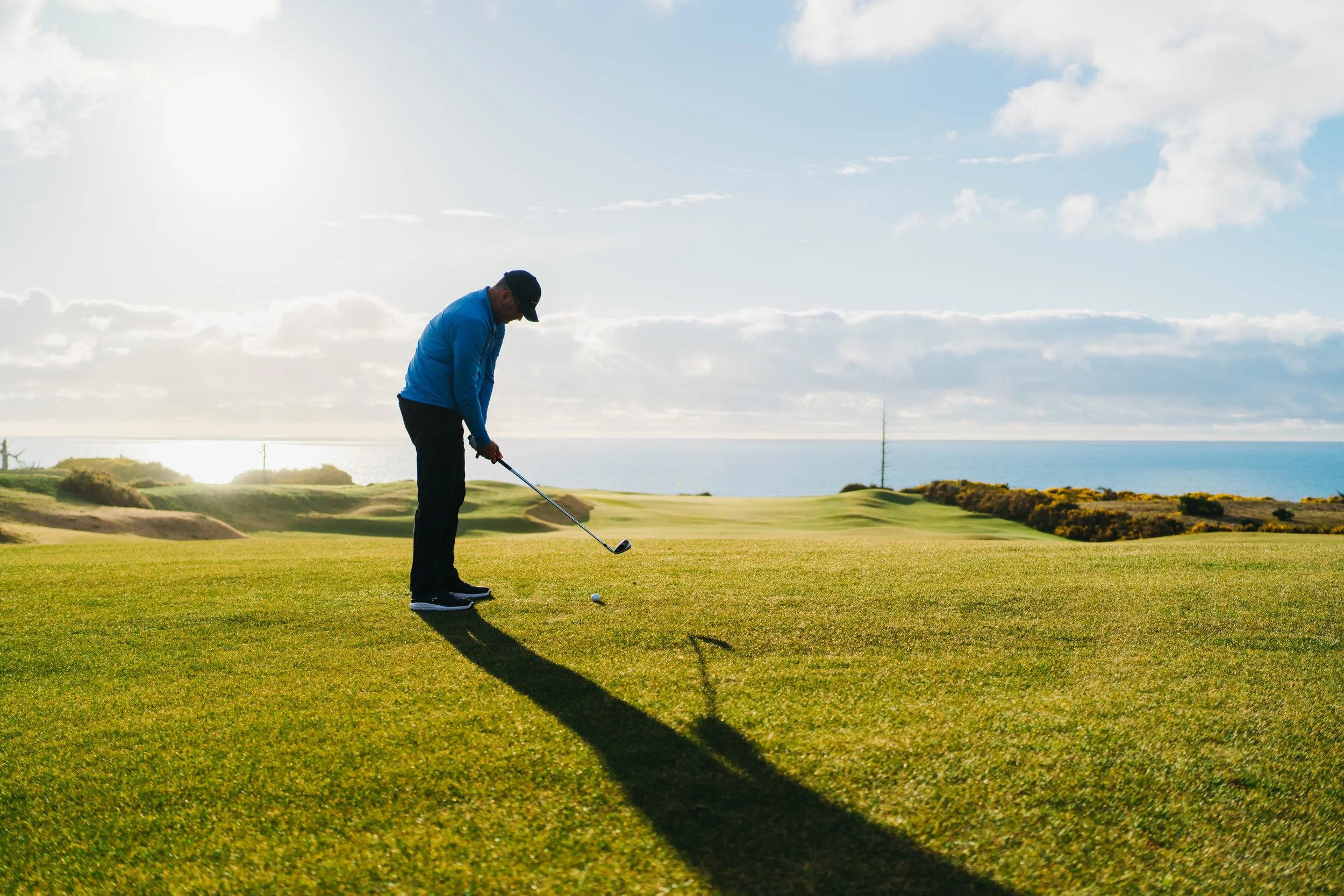 A man playing golf on a green field near the coast under a partly cloudy sky.