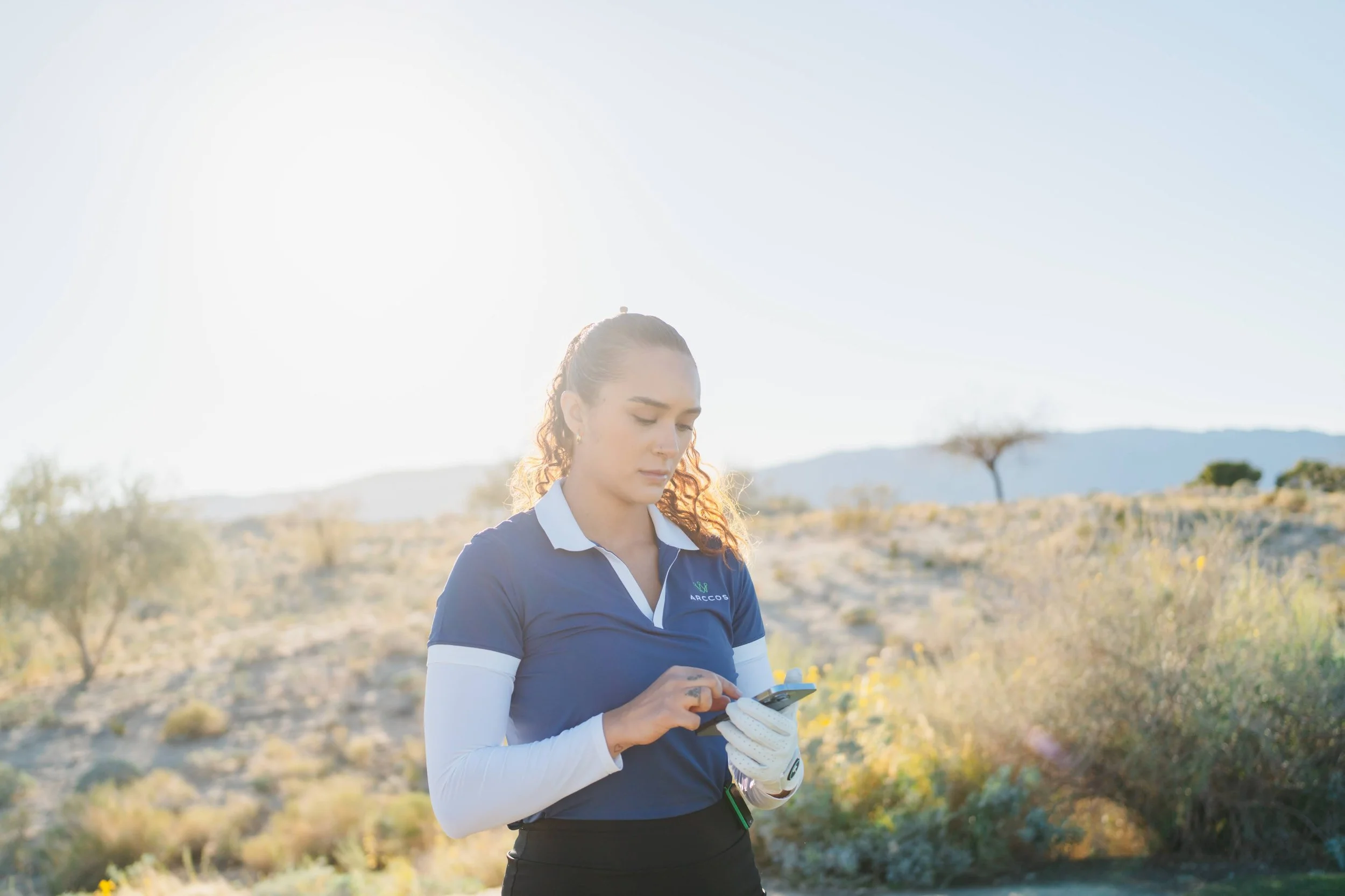 Woman in a blue sports shirt using a smartphone outdoors in a sunny desert landscape with sparse bushes and distant mountains.