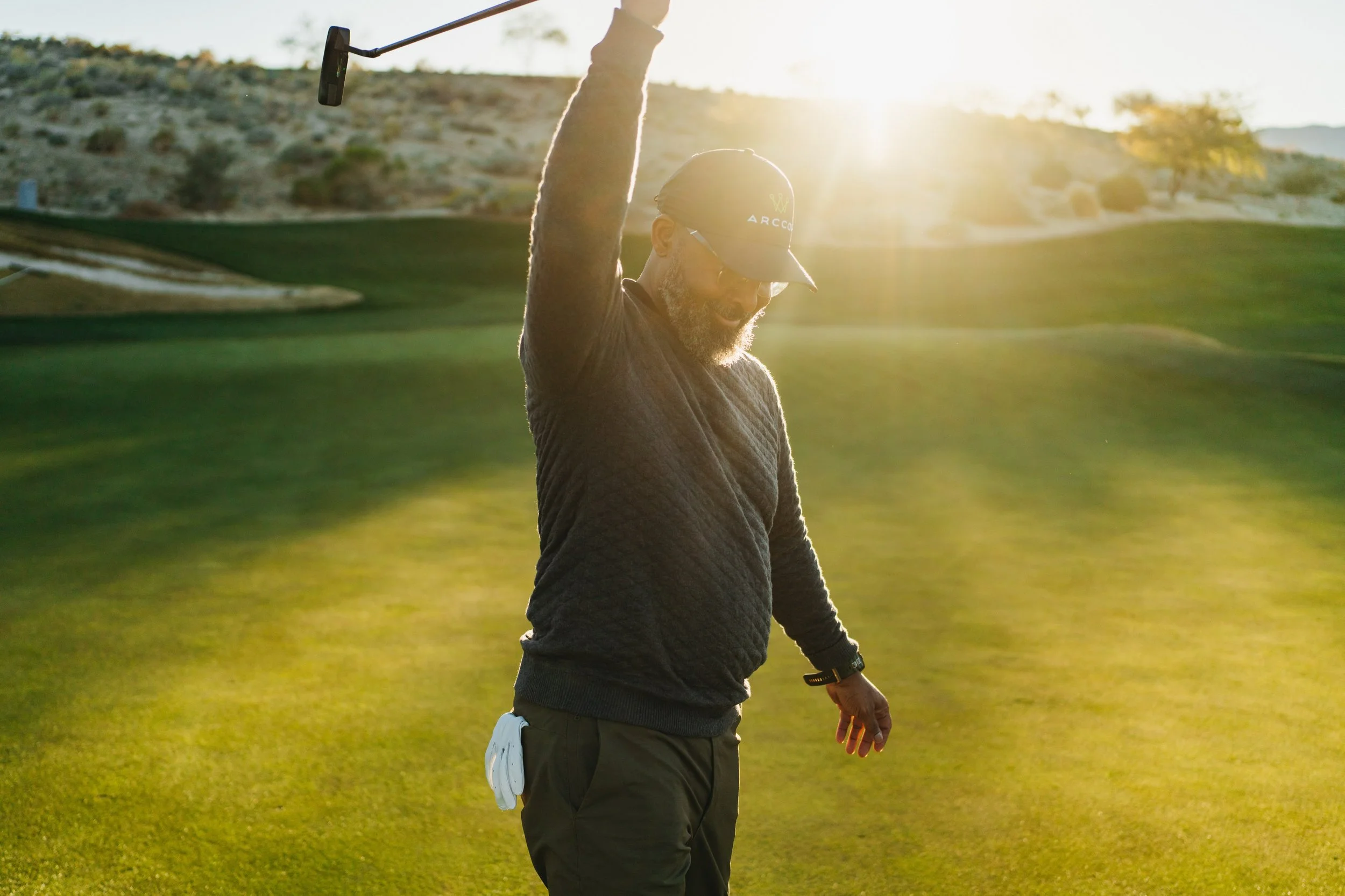 A man on a golf course raising his golf club in the air, wearing a cap, sunglasses, and a sweater, with the sun setting in the background.