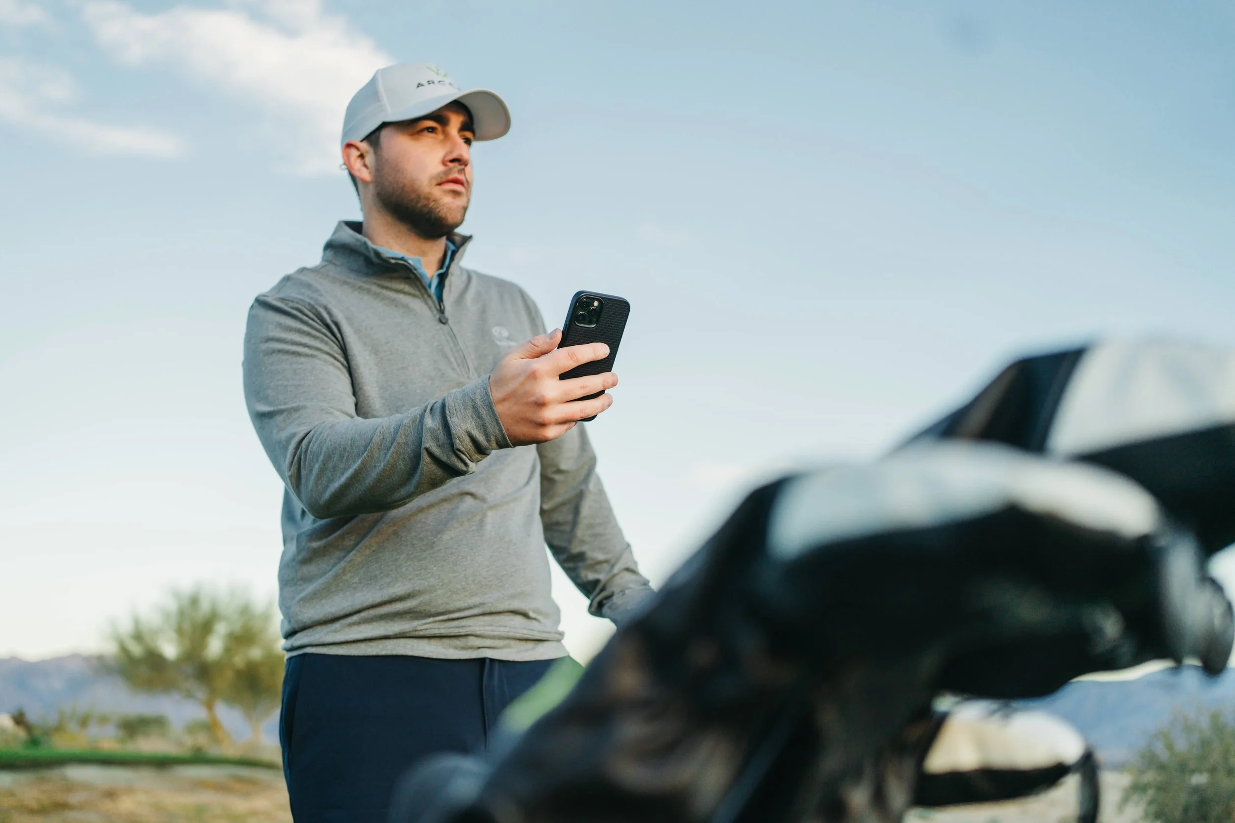 Man in gray quarter-zip jacket and white cap holding smartphone near golf clubs on golf course.
