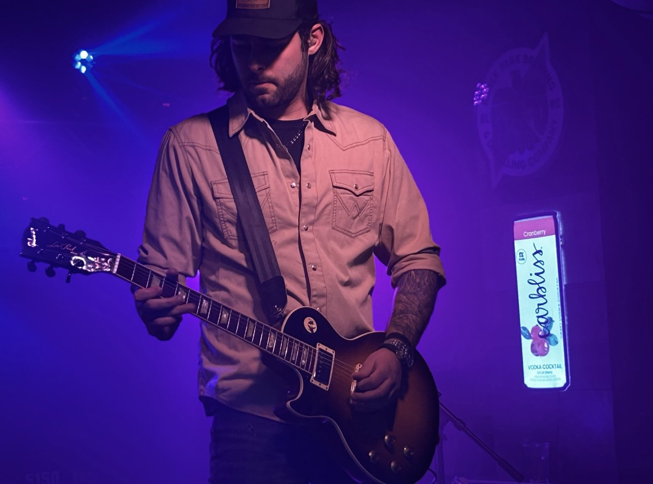 A man playing an electric guitar on stage with purple lighting, wearing a beige shirt, a black cap, and glasses, with a sign in the background advertising a cranberry vodka cocktail.