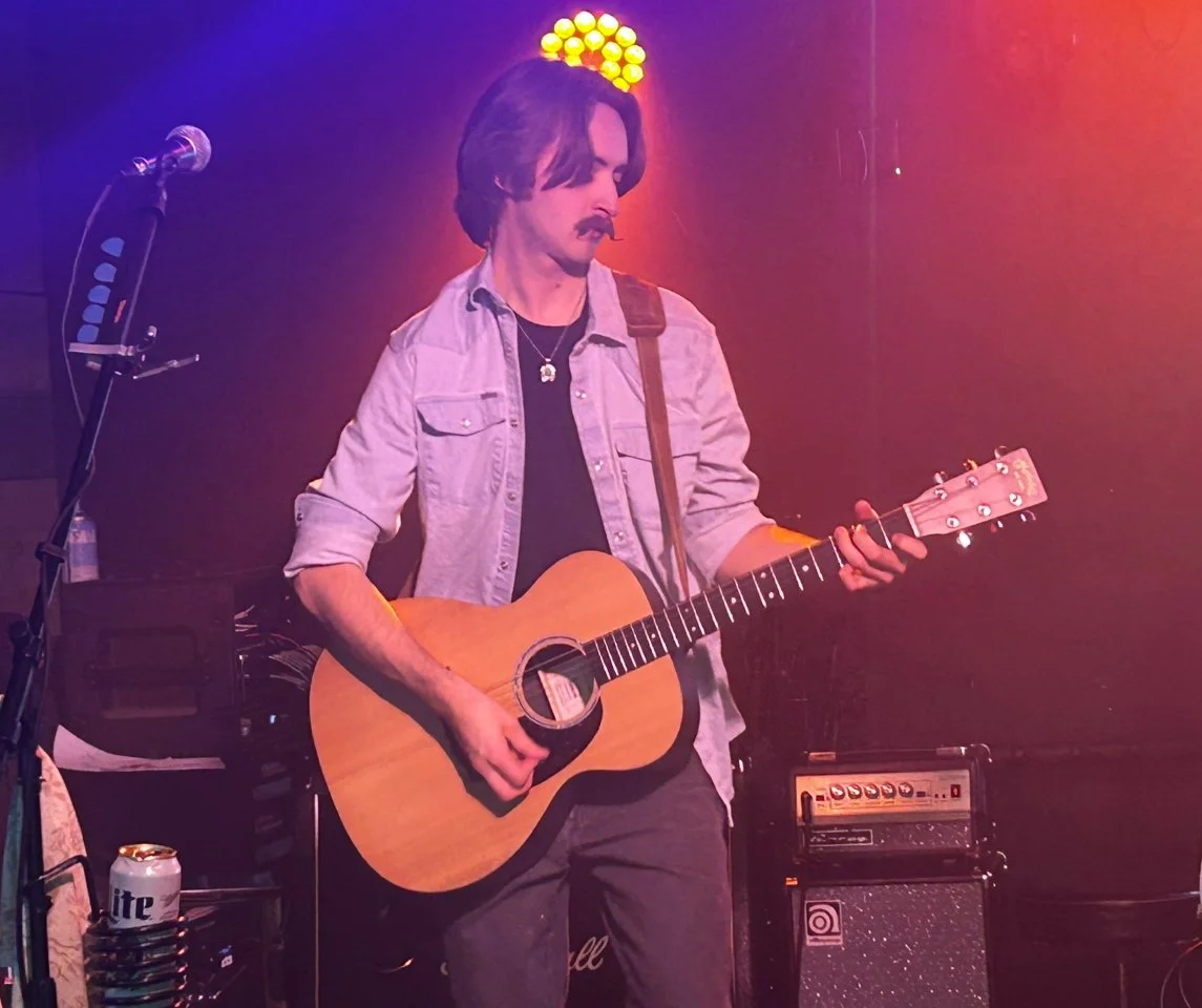 Man playing acoustic guitar on stage with colorful lights behind him.
