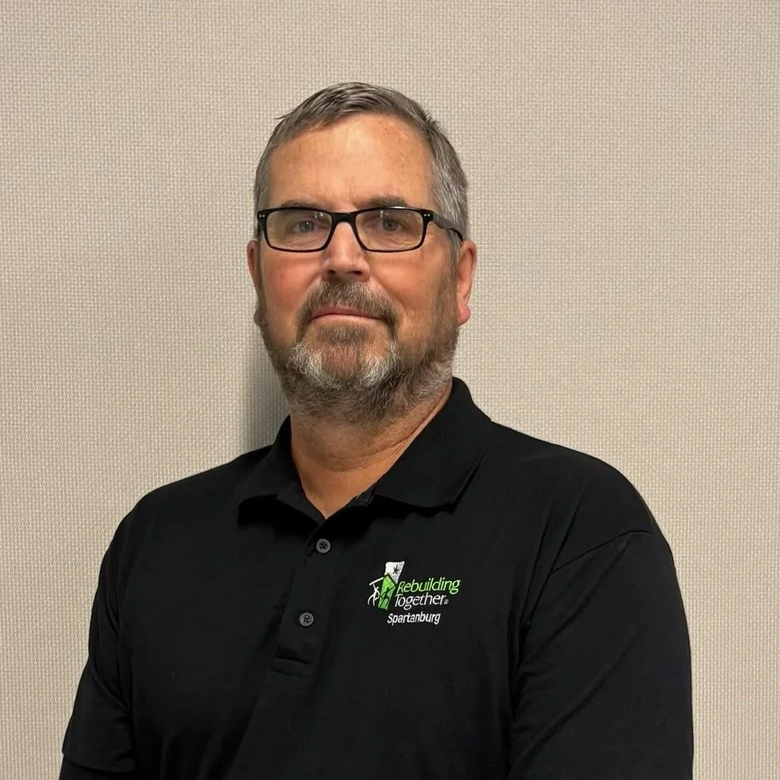 White man with a beard and glasses, standing in a black shirt with the company logo, "Rebuilding Together," on it.