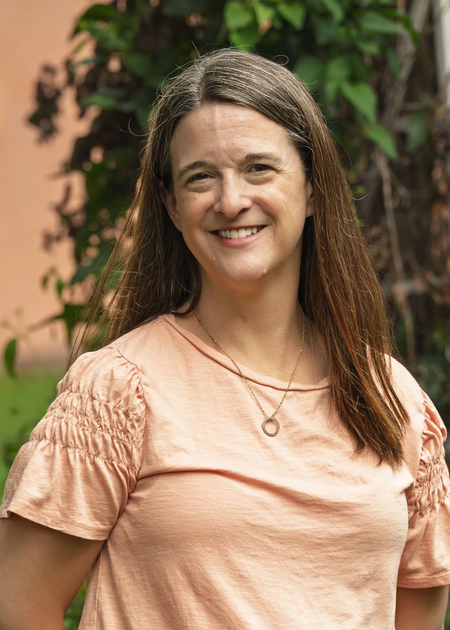 Close-up of a woman with long brown hair, smiling, wearing a peach-colored top with puffed sleeves and a silver necklace, standing outdoors with green foliage behind her.