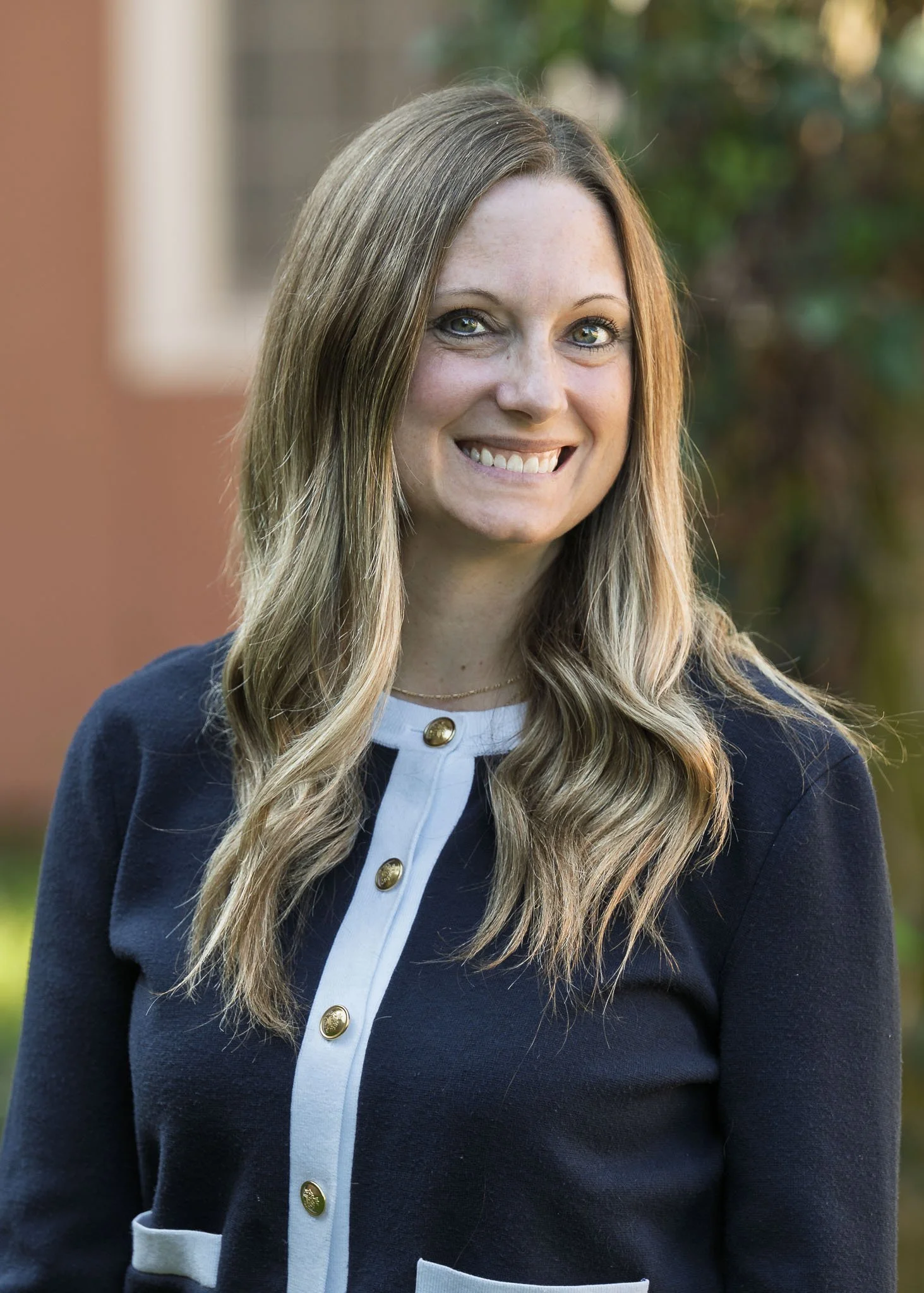 Close-up of a woman with long, wavy blonde hair, blue eyes, smiling, wearing a navy blazer with gold buttons and a white blouse with gold buttons, outdoor background with blurred greenery.