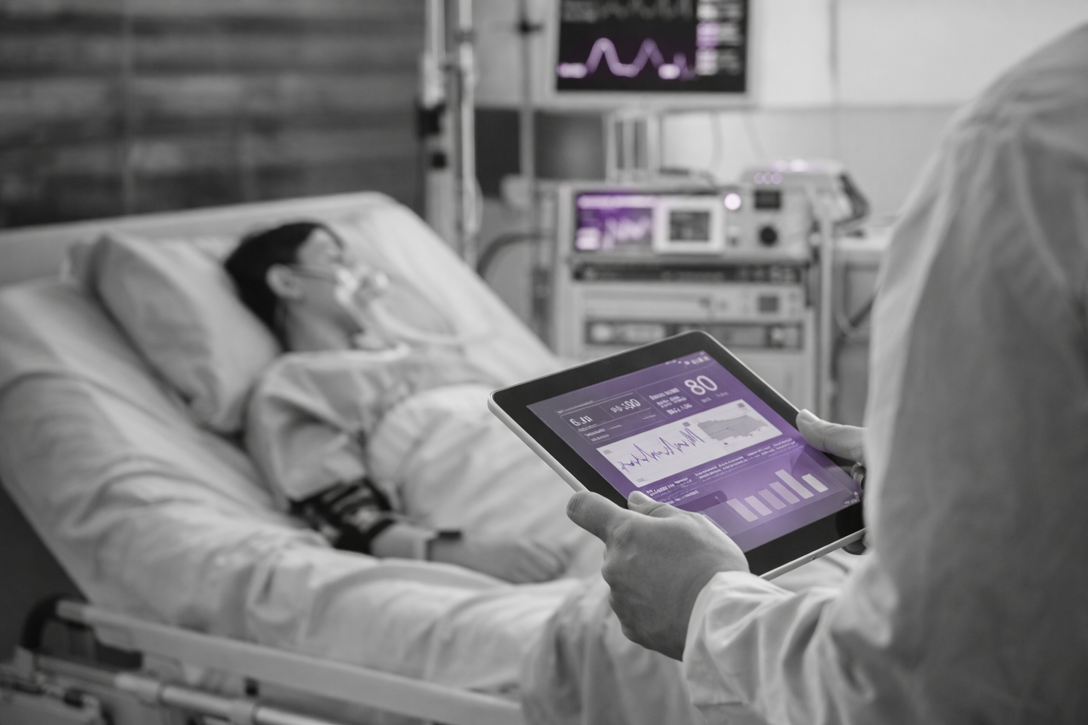 Medical professional holding a tablet displaying patient vital signs, with a patient lying in a hospital bed connected to monitoring equipment.