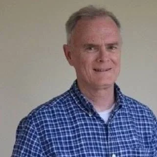 A man with short gray hair wearing a blue checkered shirt, standing against a plain wall.