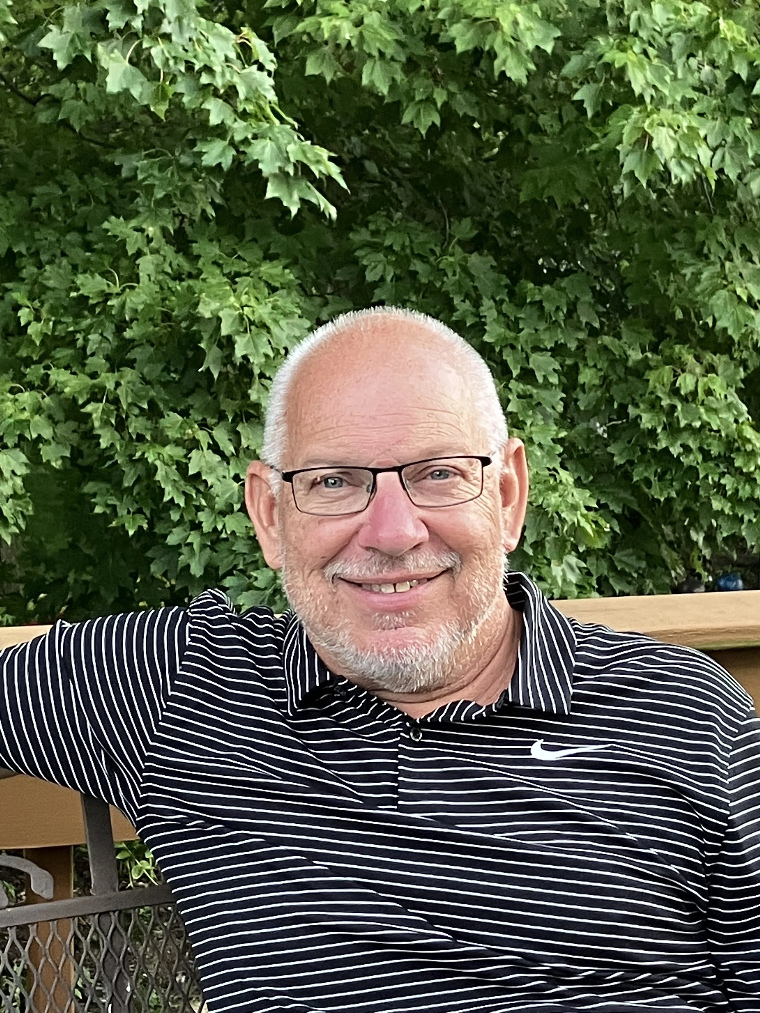 Smiling older man with glasses and a beard taking a selfie outdoors, with green leafy trees in the background.