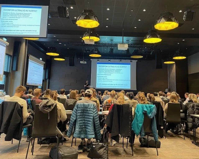 En forsamling av mennesker i et konferanserom, som deltar i en presentasjon med store skjermer som viser tekst om behandling av akutt hjertesvikt.