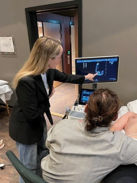 En kvinne i svart dress viser en ultralydskjerm til en pasient som ligger på sengen.