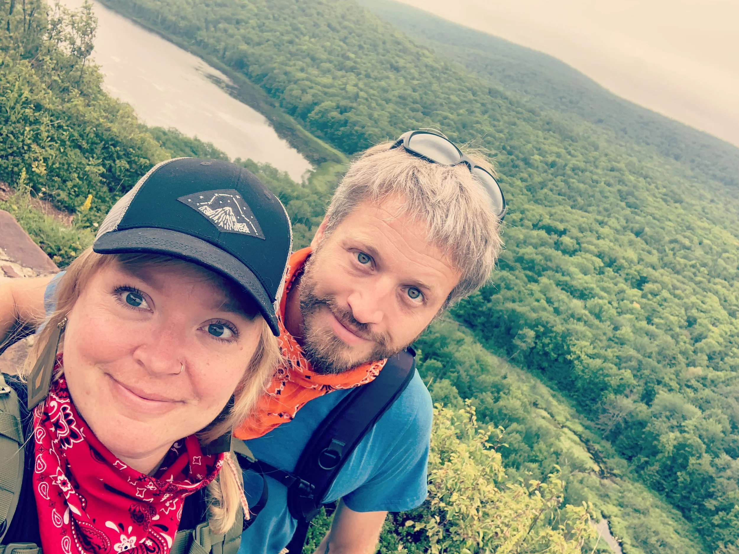 A smiling woman and man taking a selfie together on a hiking trail overlooking a lush green landscape with a river and hills.